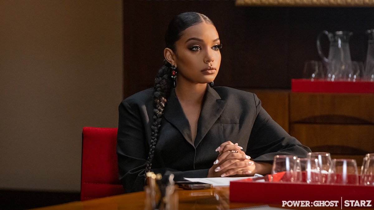 Woman with braided hair and earrings sits at a table with glasses and a notepad, wearing a black blazer.