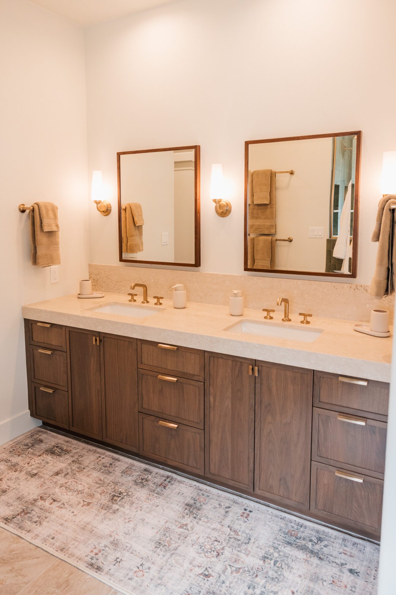 Bathroom vanity with two mirrors, two sinks, and wooden cabinets, beige countertop, and towels on wall hooks.