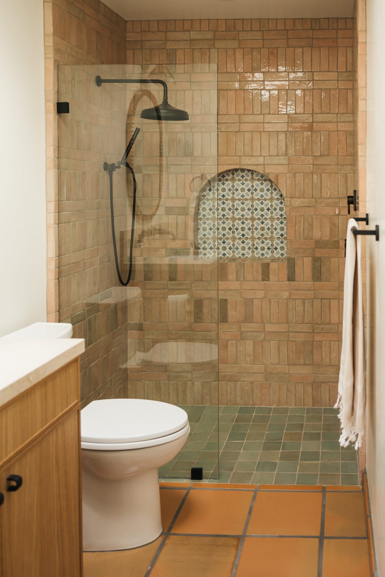 Bathroom shower area with tiled wall, small arched niche, and a showerhead, next to a toilet and wooden cabinet.