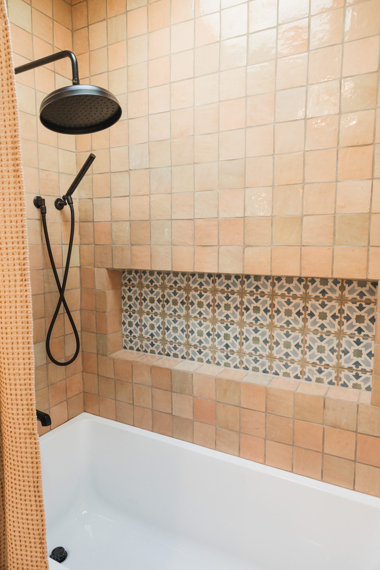 Shower area with black showerhead, beige tiled wall, and decorative tile niche, with a white bathtub below.