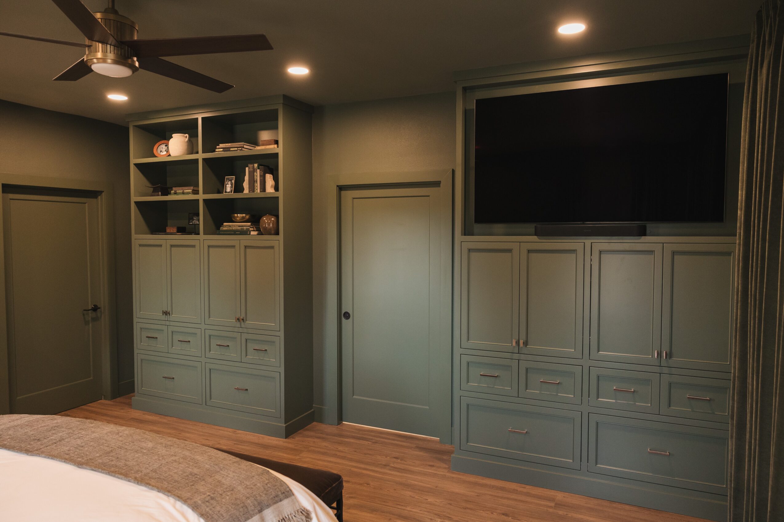 Bedroom corner with built-in cabinets, shelves, a door, and a large TV, with a ceiling fan and wooden floor.