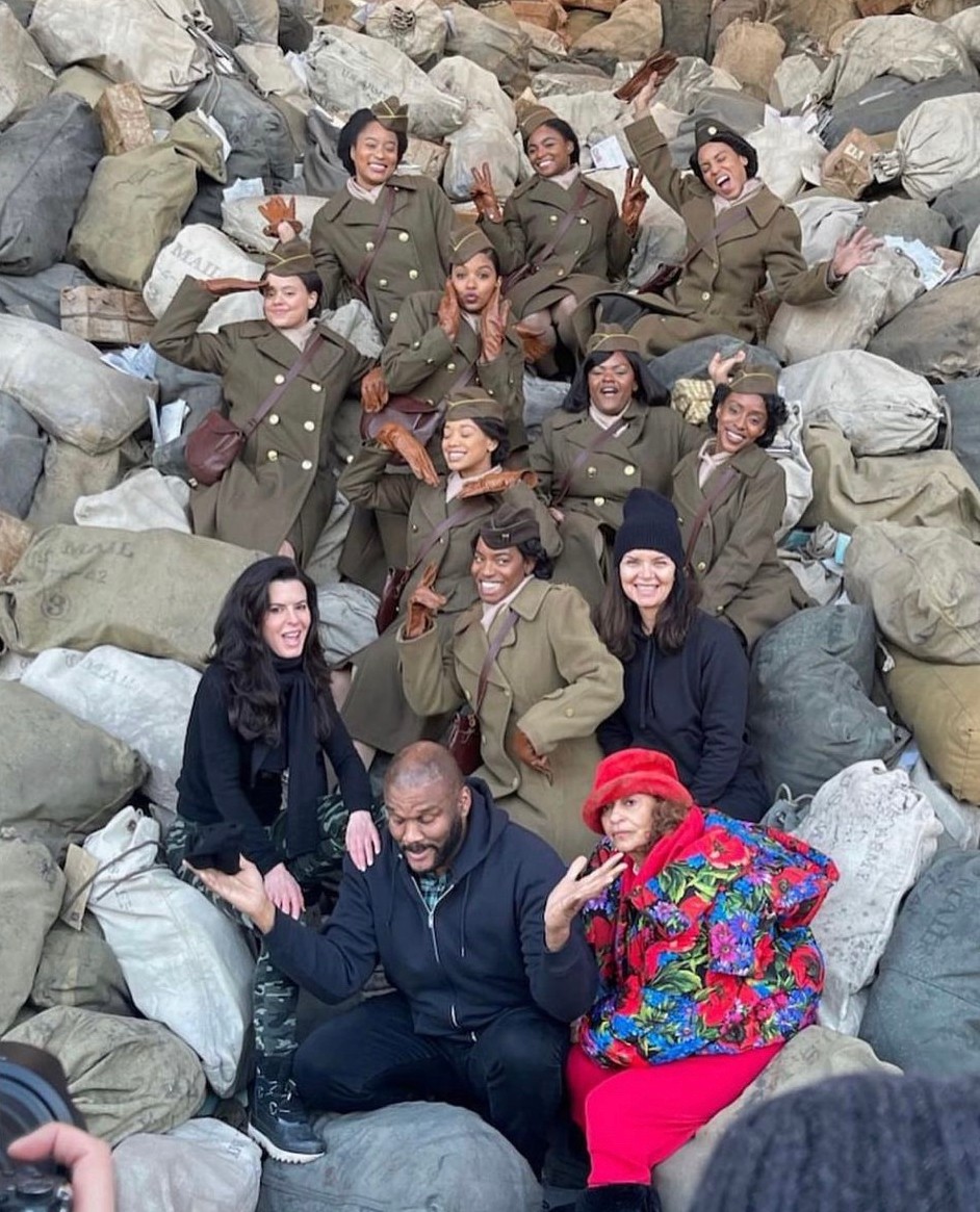 Group of people in military uniforms and colorful clothing sitting among rocks and bags outdoors.