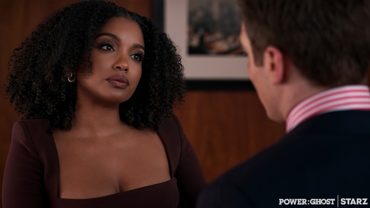 A woman with curly hair looks at a man with short hair in a room with framed pictures, serious expression.