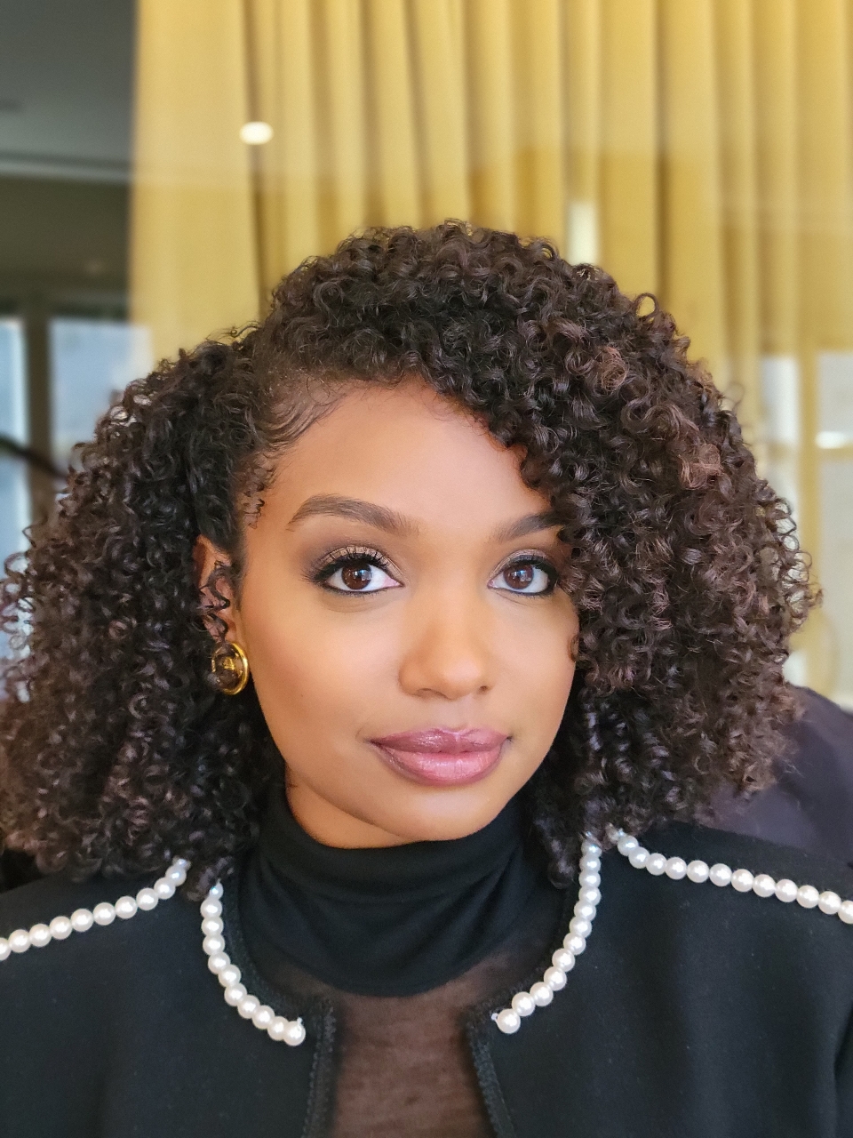 Portrait of a woman with curly hair, wearing a black top, pearl necklace, and gold earrings, in front of yellow curtains.