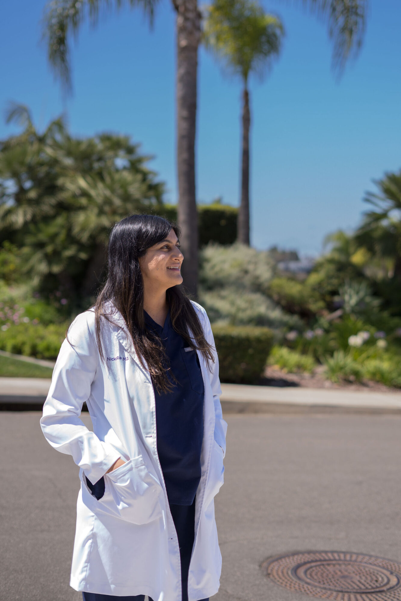Woman in white coat standing outdoors with palm trees and greenery in background, smiling and looking to the side.