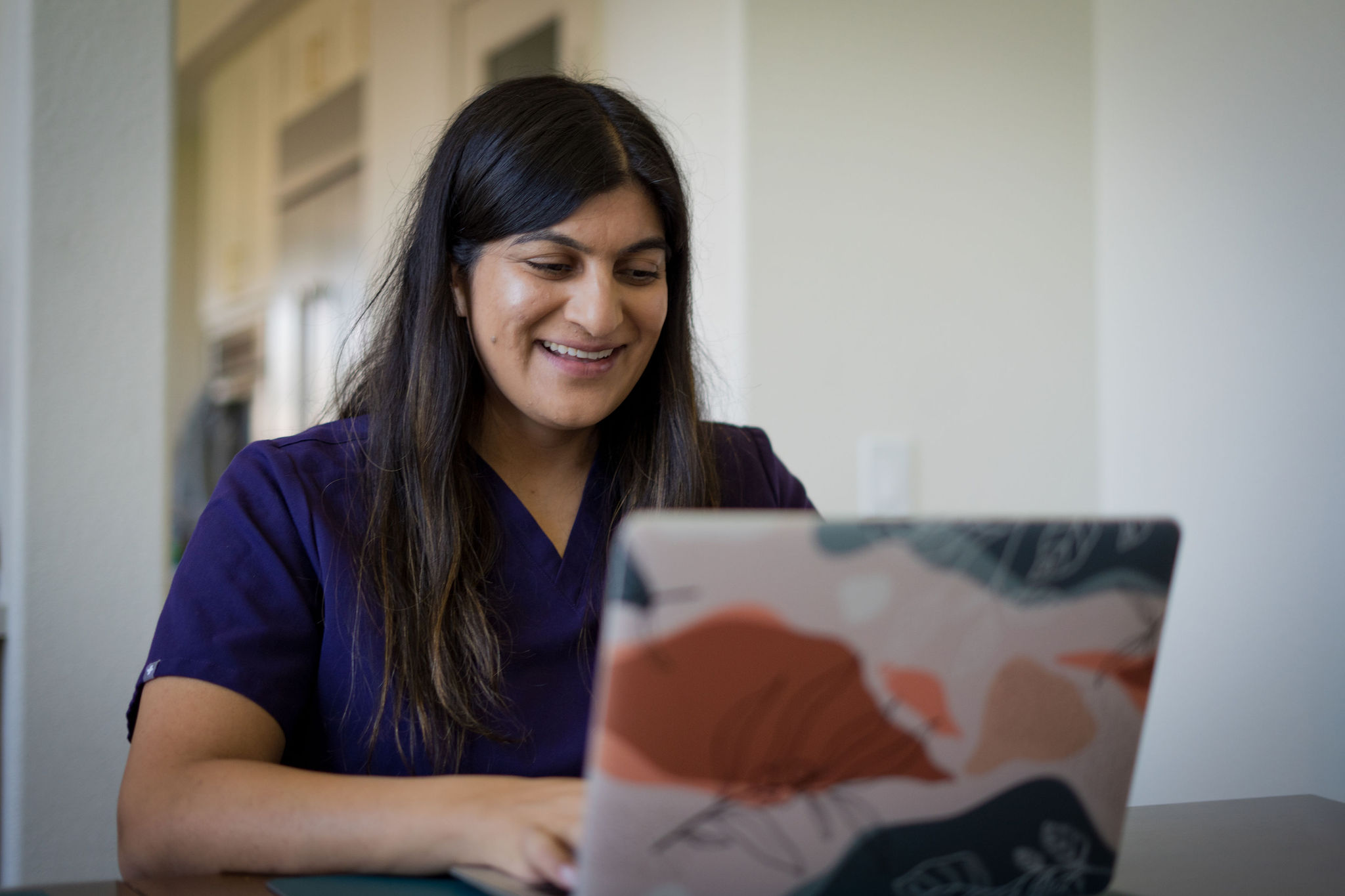 Woman with long dark hair smiling while looking at a laptop, wearing a dark purple top, in a bright room.