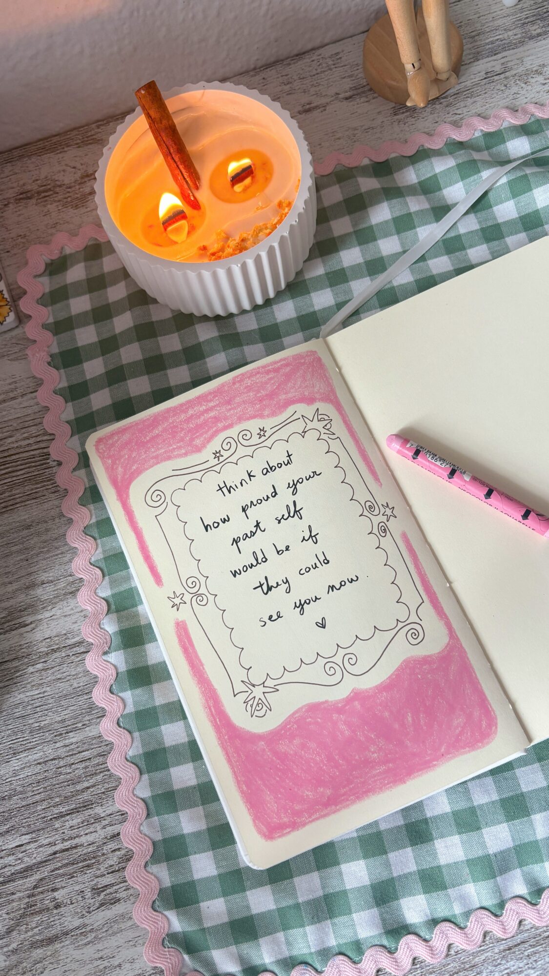 Open notebook with handwritten note, pink border, pink pen, candle, and notebook on checkered tablecloth.