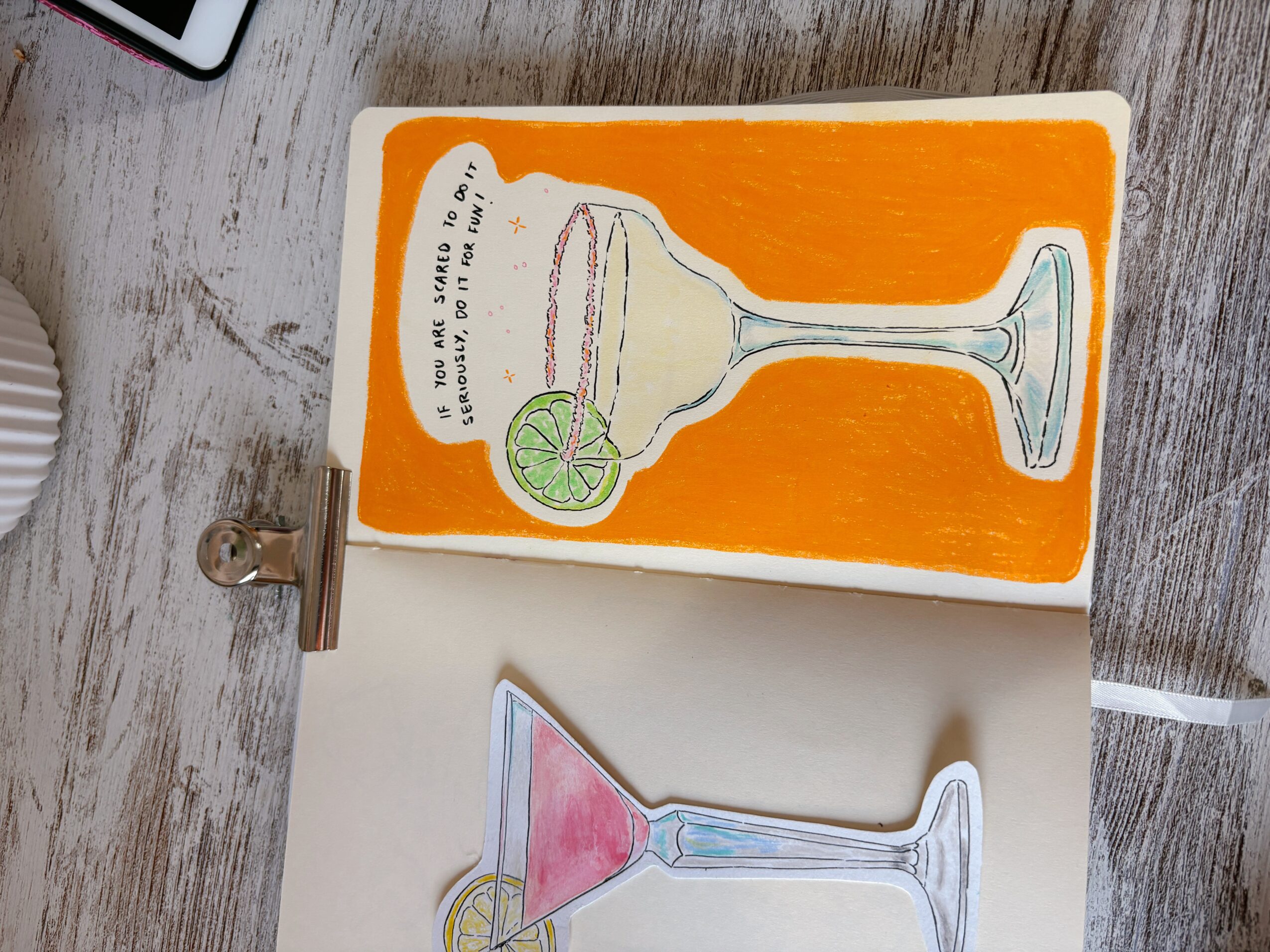 Open notebook with colorful drawing of a cocktail glass with a lime slice on a wooden surface.