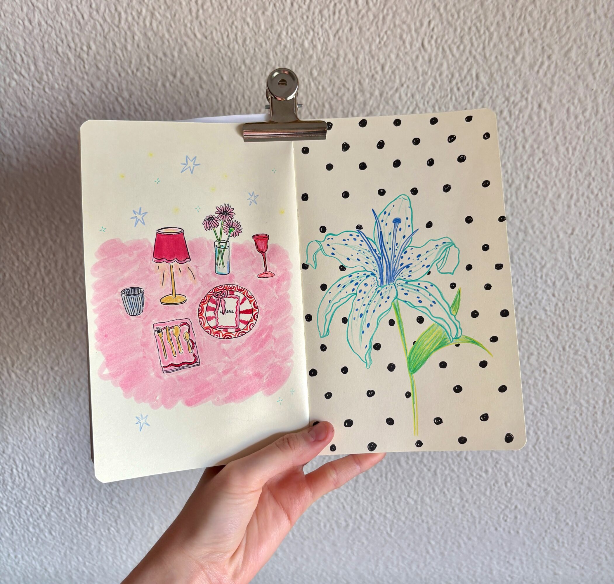 Open sketchbook with colorful drawings of lamps, a clock, and a flower on polka dot background, held against a wall.