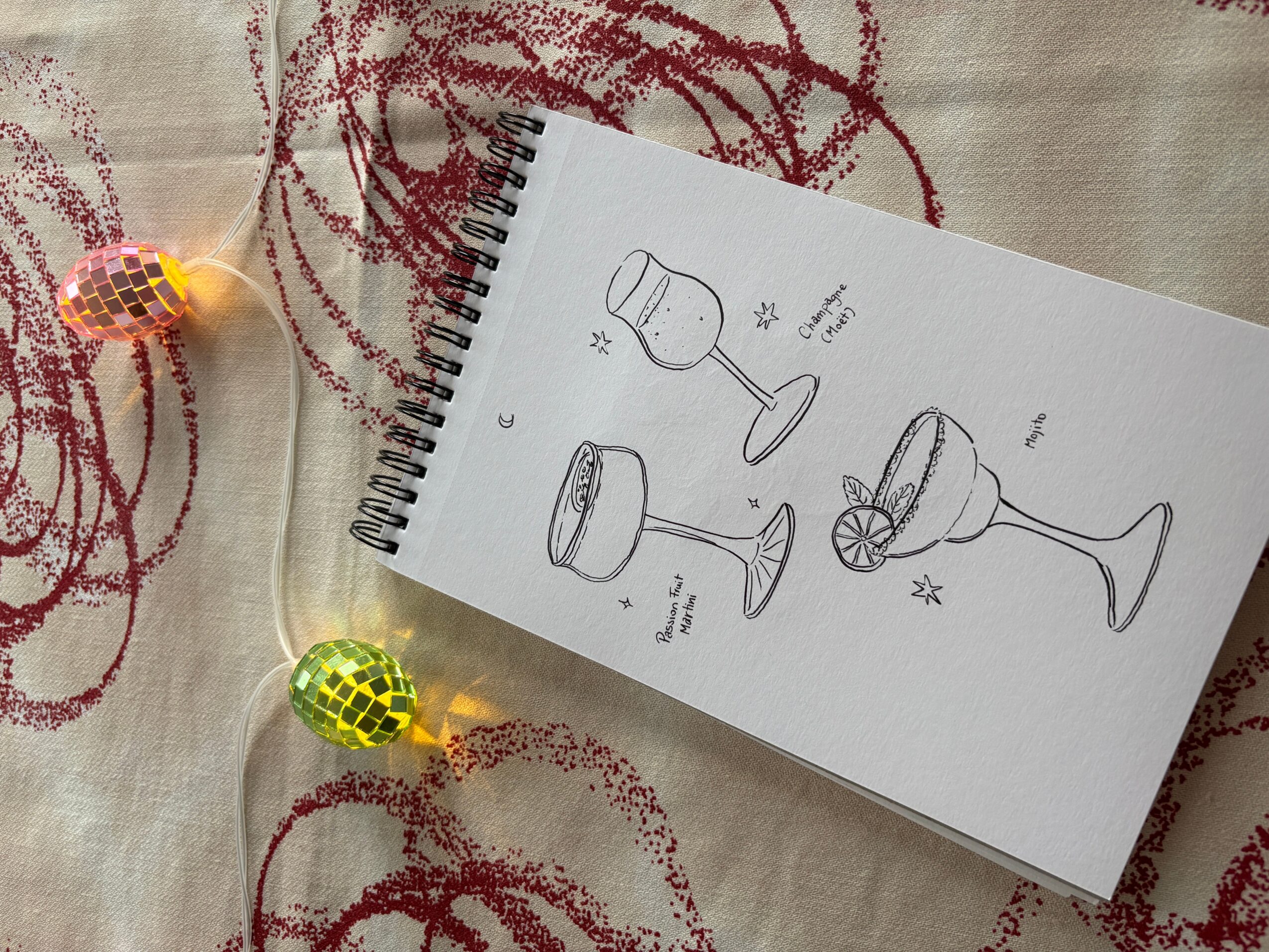 Sketch of three cocktail glasses with labels and decorative elements on a notebook page, placed on fabric with string lights nearby.