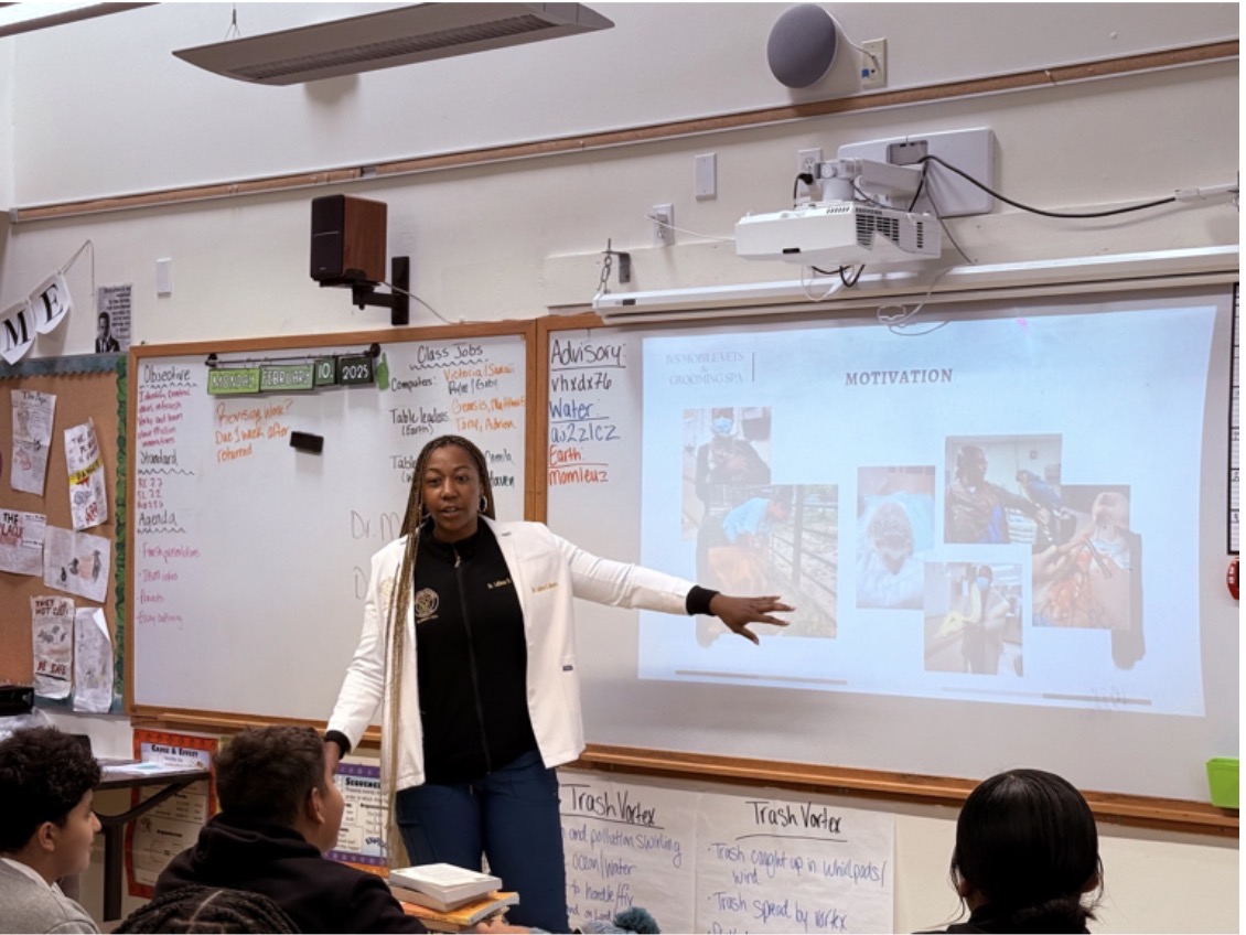 Teacher standing in front of a classroom, pointing at a presentation on a whiteboard with students seated and listening.