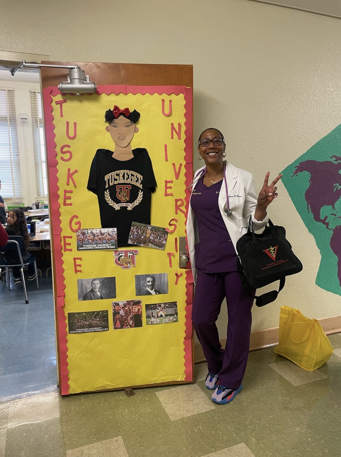 Woman standing next to a yellow display board with photos and decorations, smiling and making a peace sign, in a school hallway.