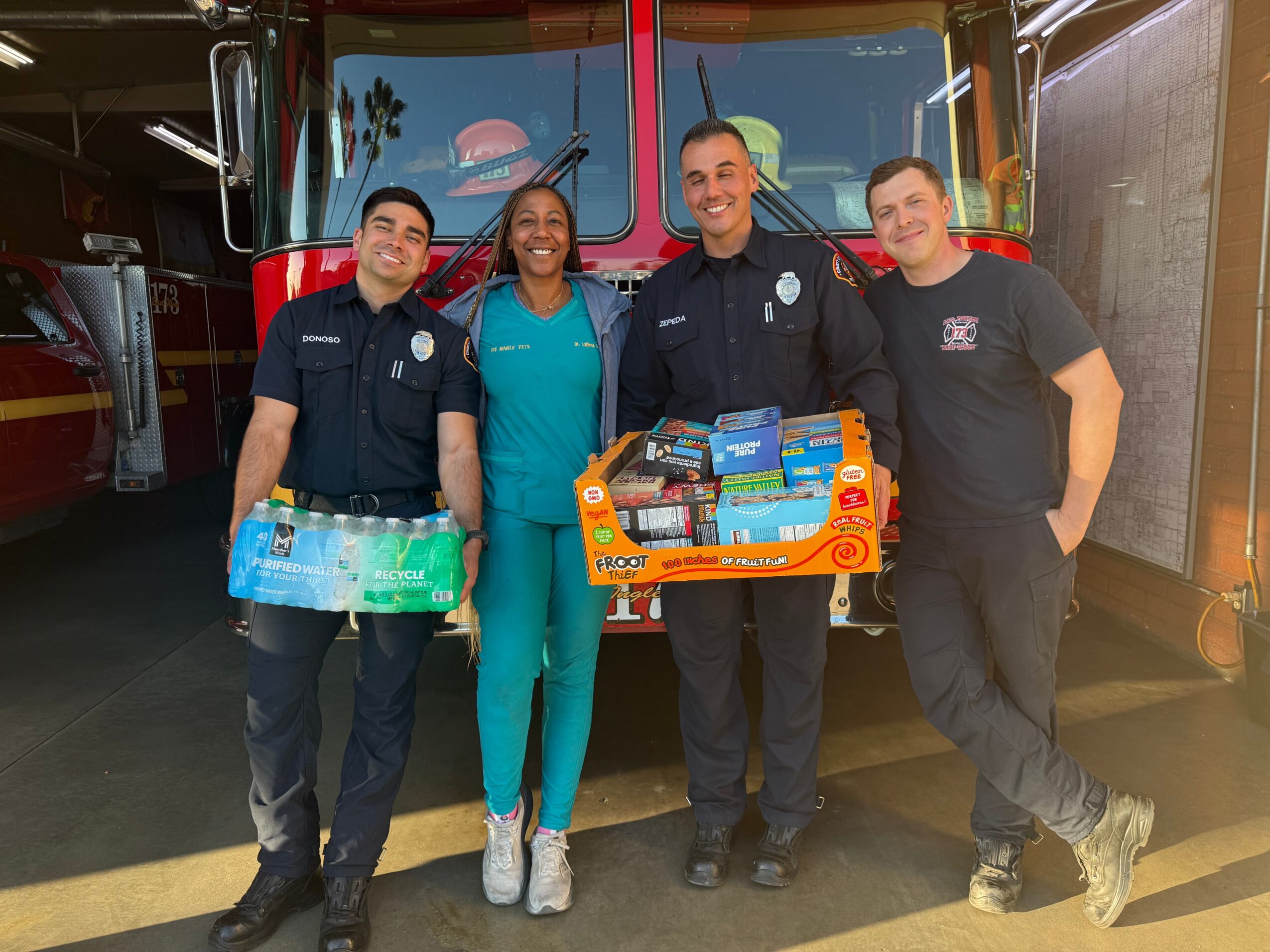 Four people, including firefighters and a woman in medical scrubs, stand in front of a fire truck holding supplies.