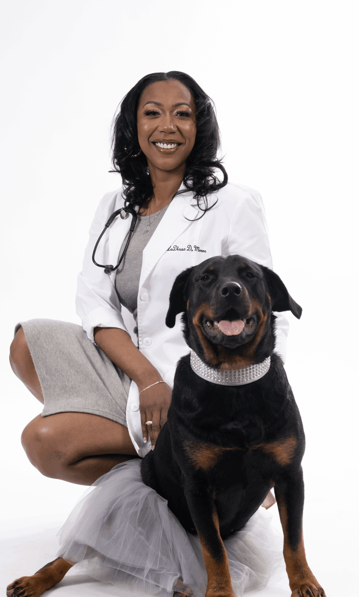 Female veterinarian in white coat kneeling with a smiling black and tan dog against a white background.