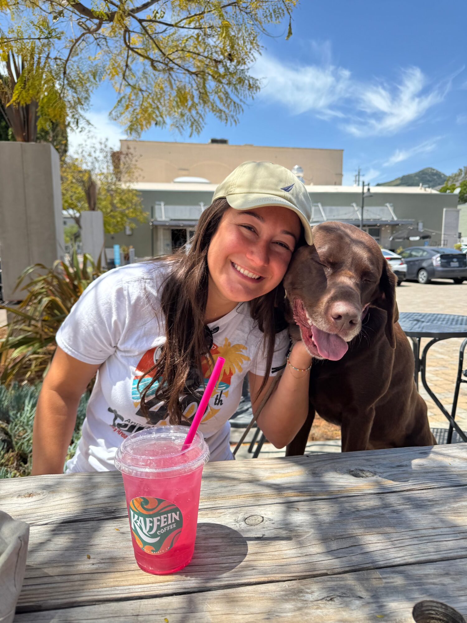 Two women sitting at a wooden table outdoors, smiling, with a pink drink and a dog on the table, under a blue sky.