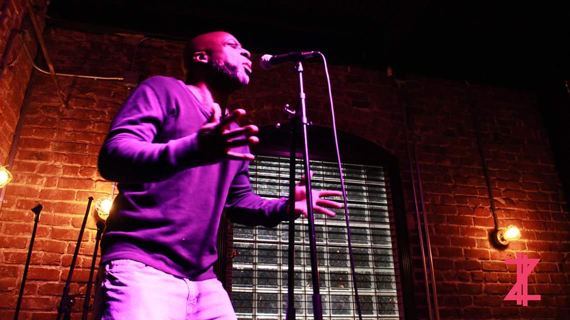 Person performing on stage with brick wall background, microphone, and stage lighting, holding a glass in one hand.