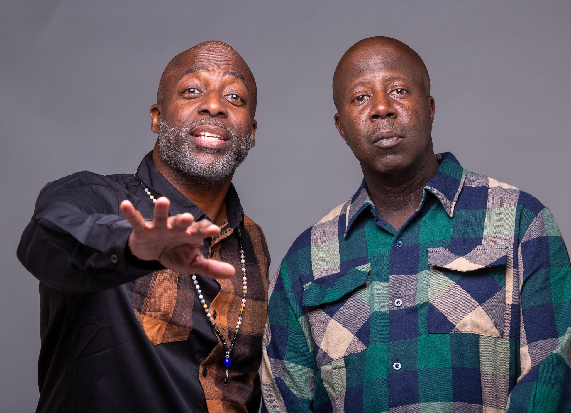 Two men stand side by side against a gray background, one gesturing with his hand, both looking at the camera.