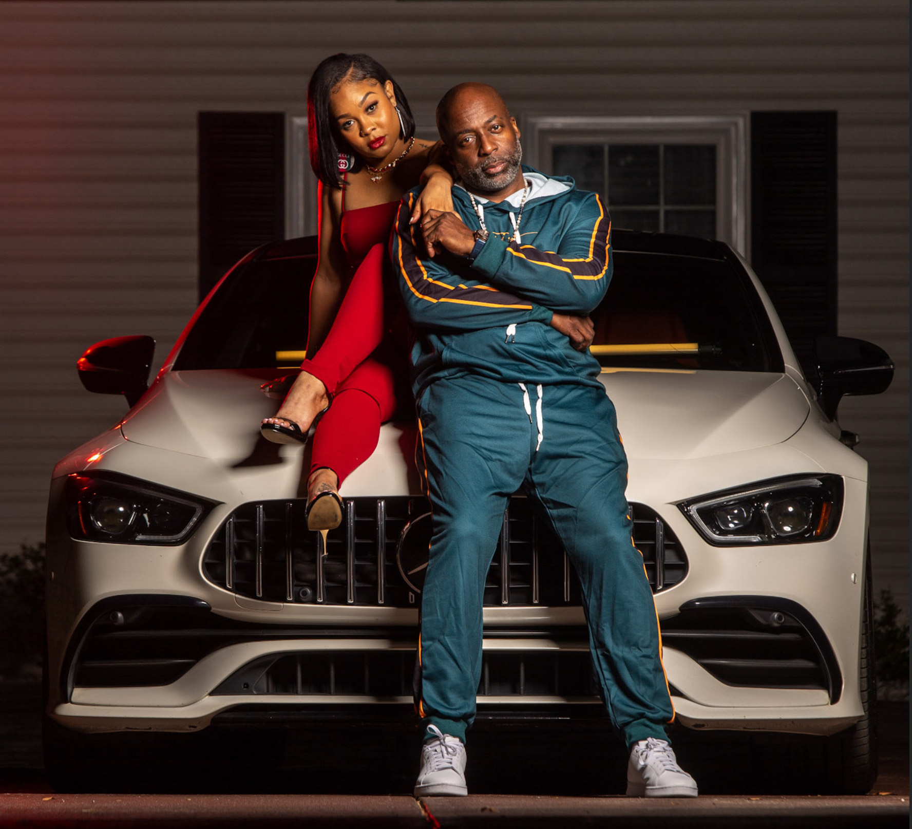 A man and woman pose in front of a white car at night, woman sitting on the hood, man standing with arms crossed.