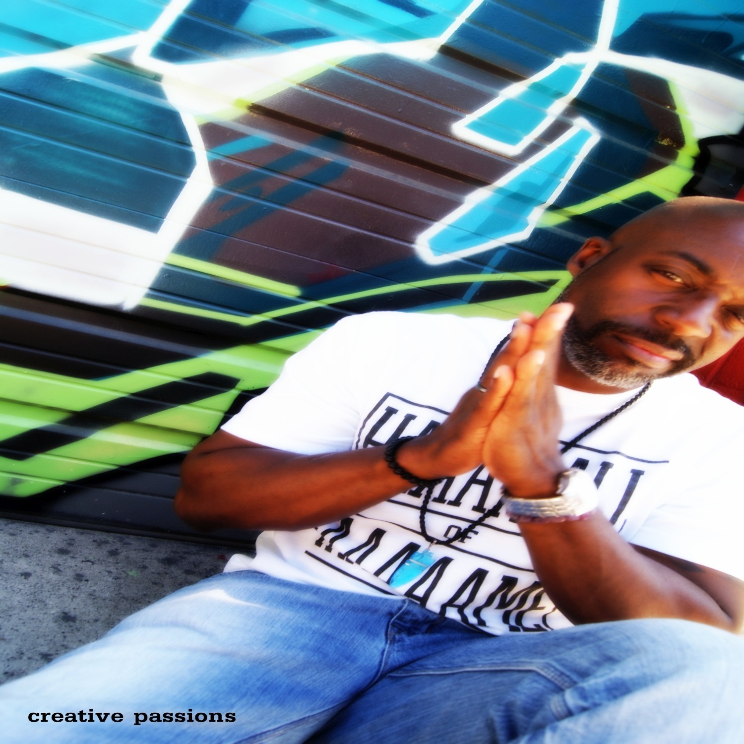Man with a beard and white T-shirt sitting with hands together in front of a graffiti wall, smiling.