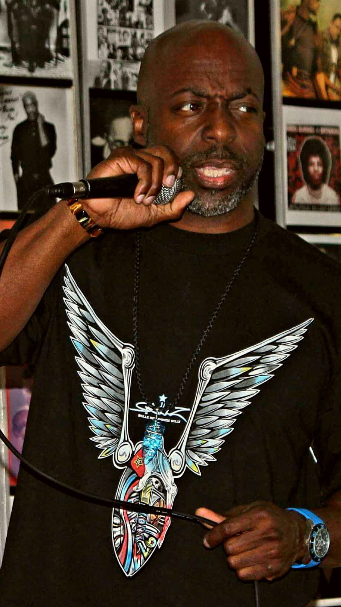 Man with a beard and bald head talking on a phone, wearing a black T-shirt with a large winged skull design, in a room with photos on the wall.