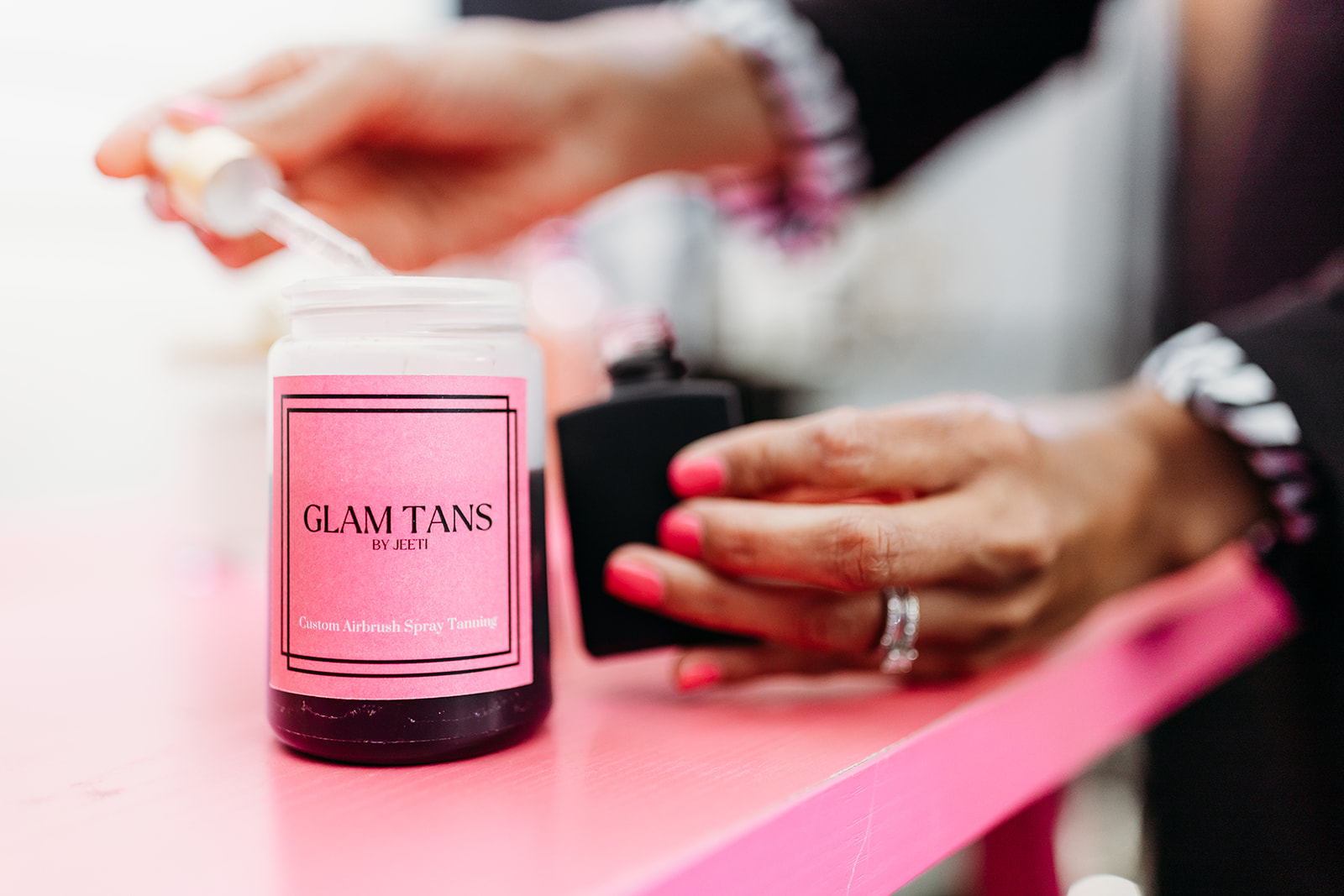 Person holding a black container with pink nail polish on a pink surface, with a pink container labeled 'GLAM TANS' nearby.