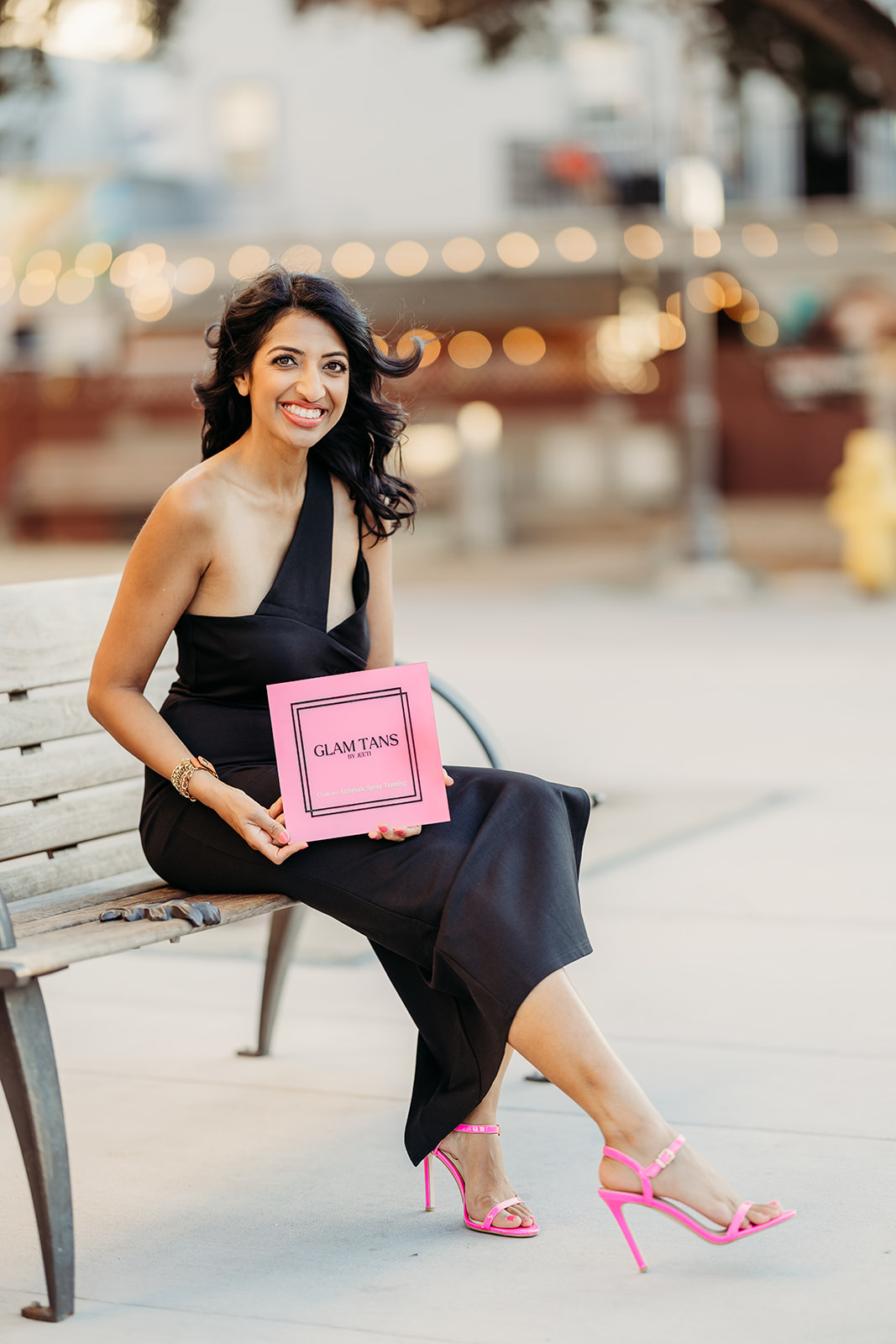 Woman sitting on a bench holding a pink sign, smiling, wearing a black dress and pink high heels, outdoors.