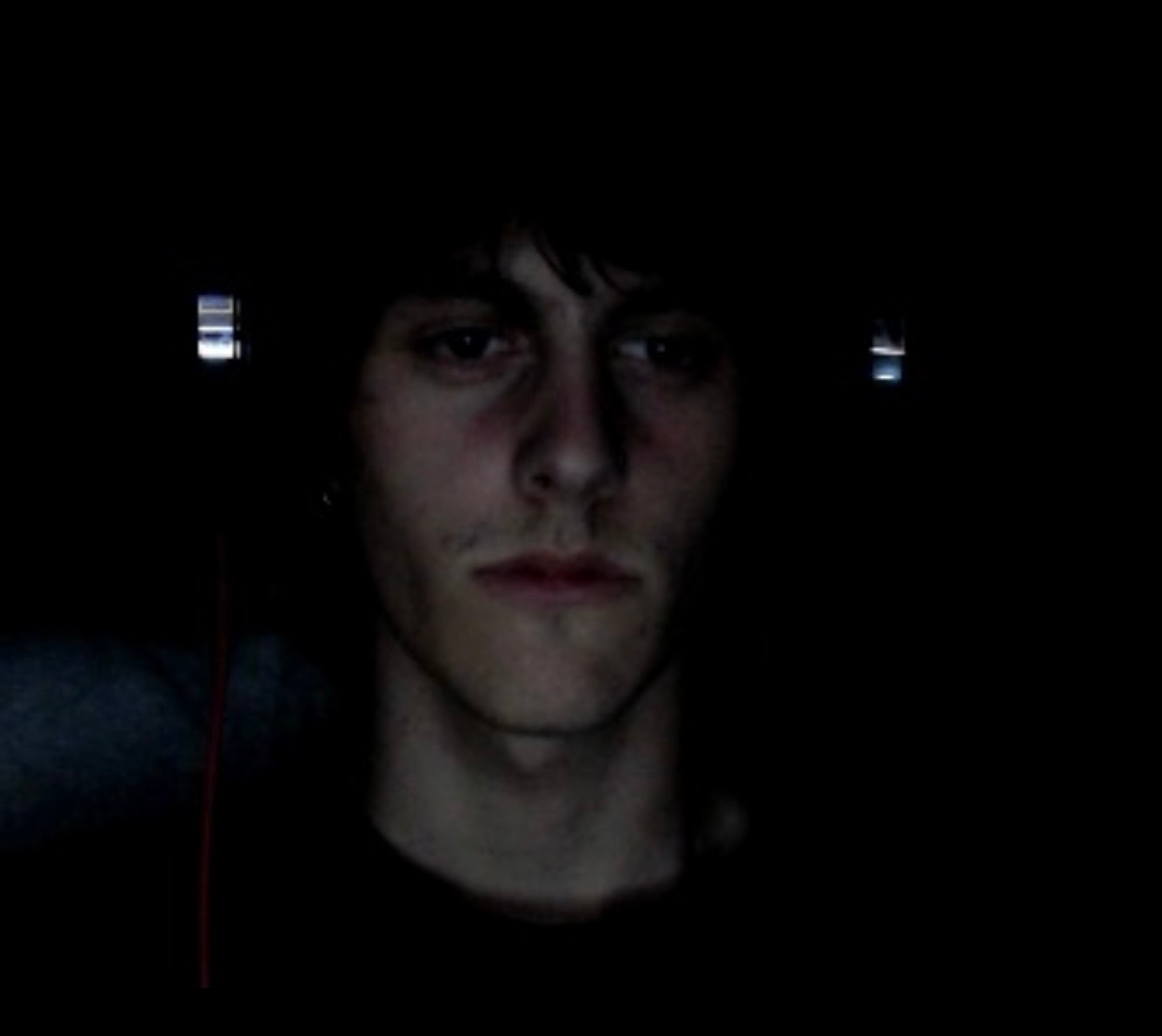 Young person with dark hair and headphones in a dark environment, face partially illuminated, serious expression.