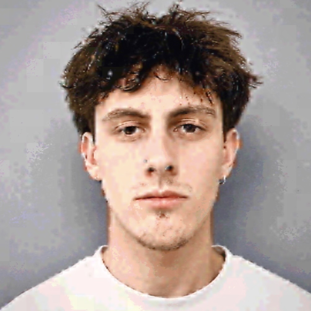 Young man with curly hair and a neutral expression, wearing a white shirt, facing forward against a gray background.