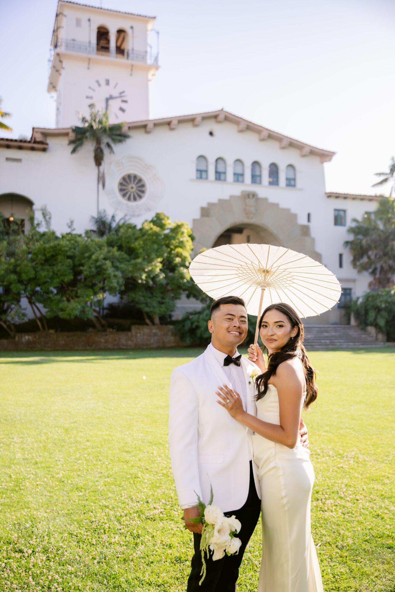A couple dressed in formal attire standing on a lawn with a white building and a parasol in the background.