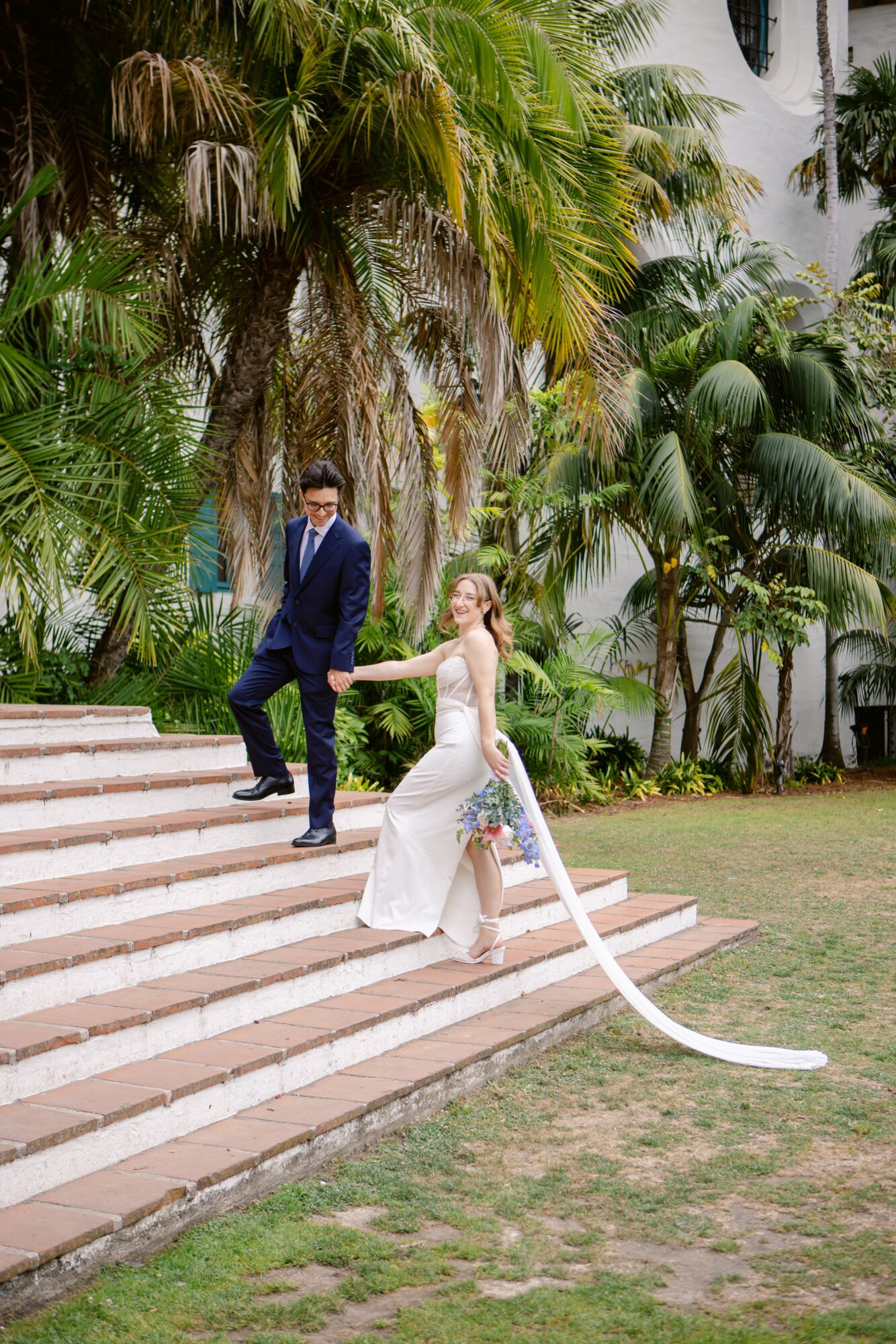 A couple in wedding attire on outdoor stairs surrounded by lush greenery and palm trees.