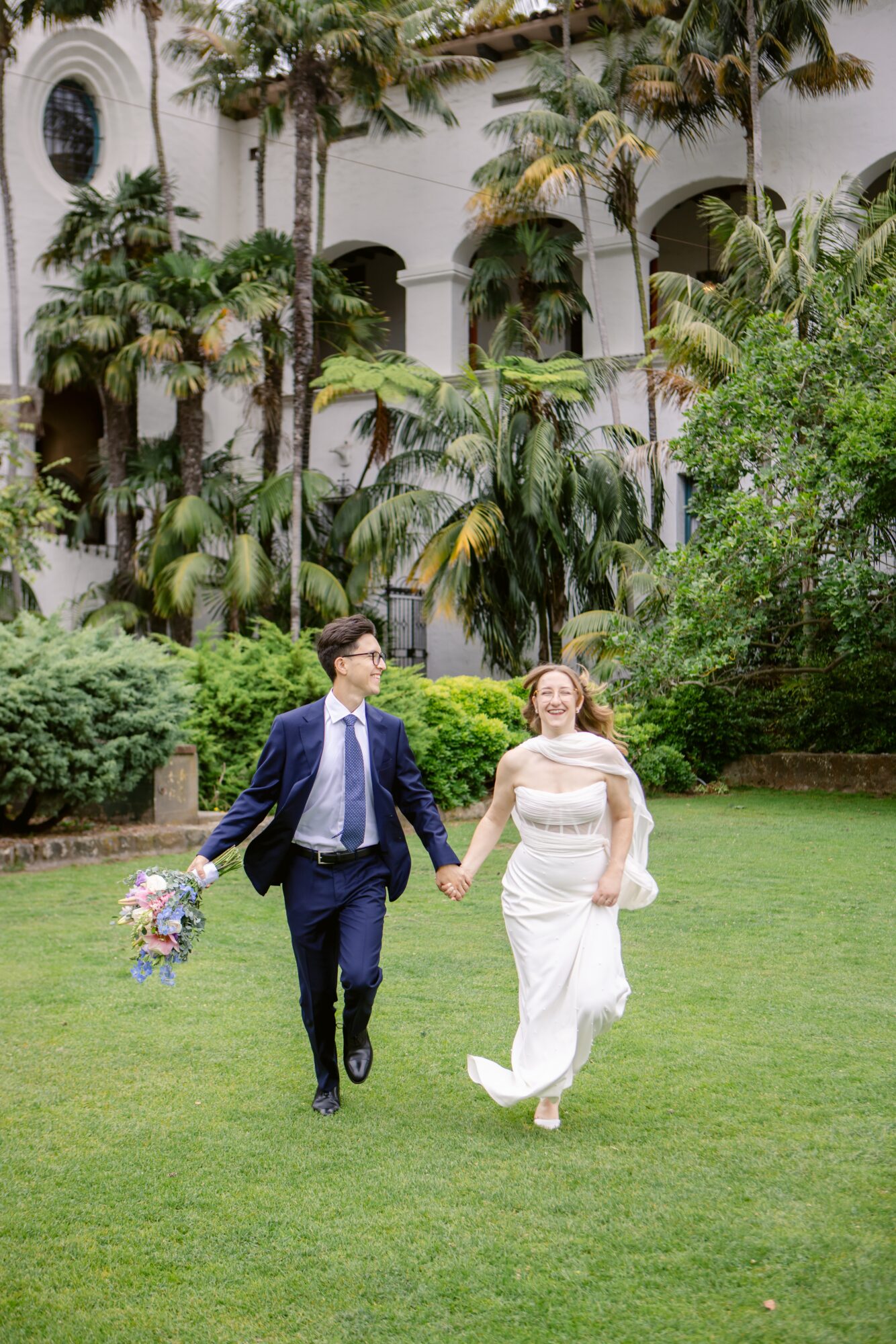 A couple holding hands, walking on grass, with a white building and lush greenery in the background.