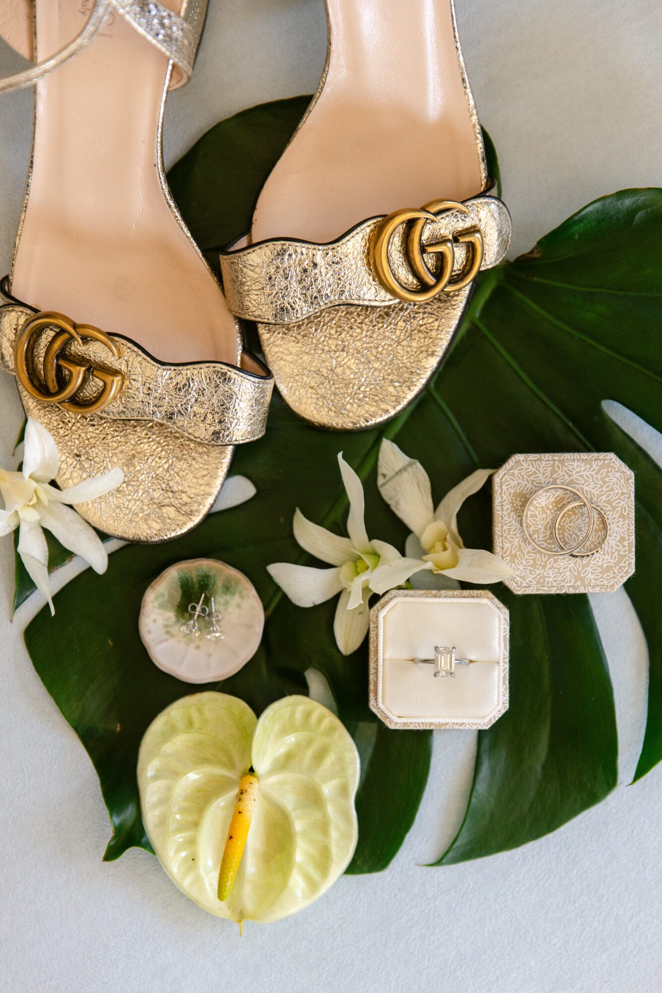 Pair of glittery beige high heels with gold buckles, surrounded by white flowers, a small box, and a decorative apple on green leaves.