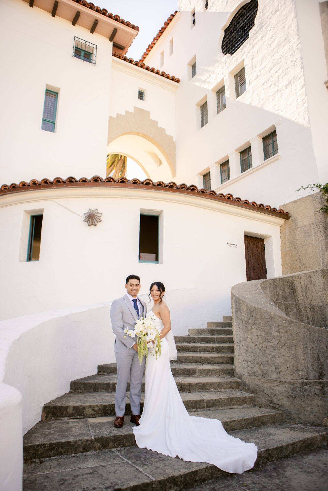 A bride and groom stand on stairs outside a white building with architectural details, sunlight illuminating the scene.