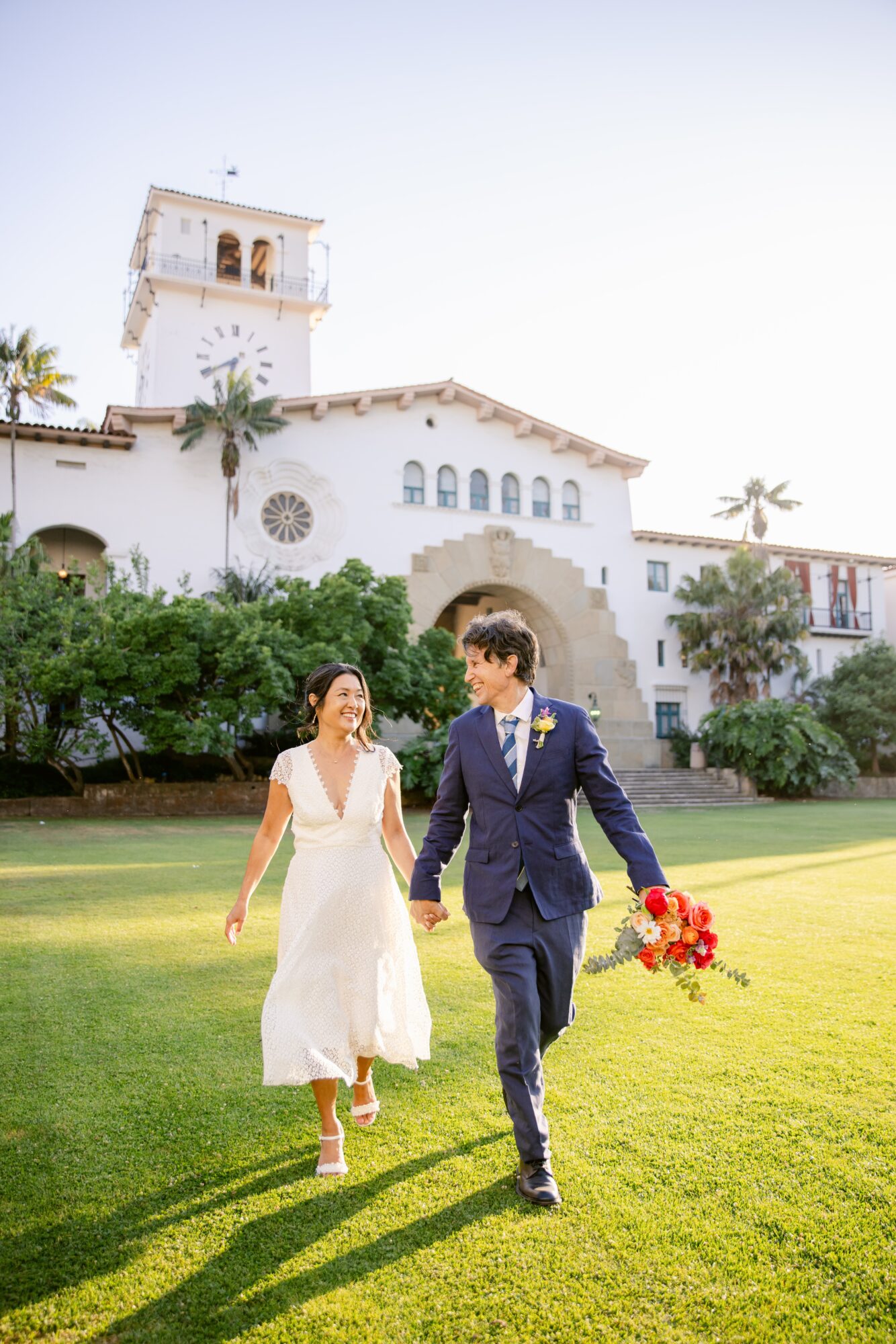 A couple walking on grass in front of a large white building with a tower, holding a bouquet of red flowers.