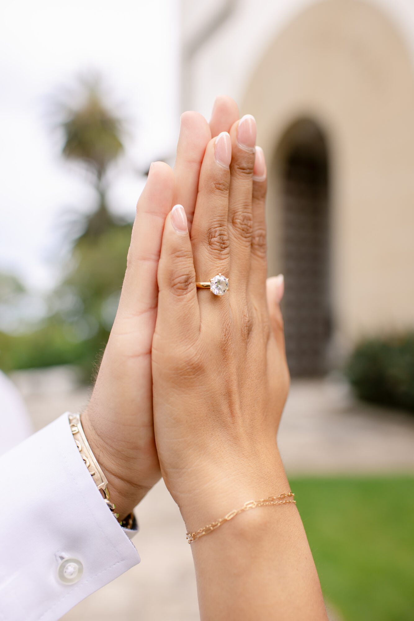 Two hands with a ring and bracelet, raised together outdoors, with a building and greenery in the background.