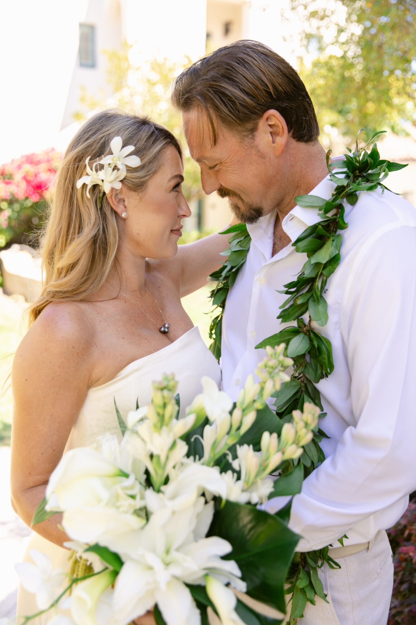 A couple on their wedding day, close together, with flowers and greenery, outdoors with trees and buildings in background.