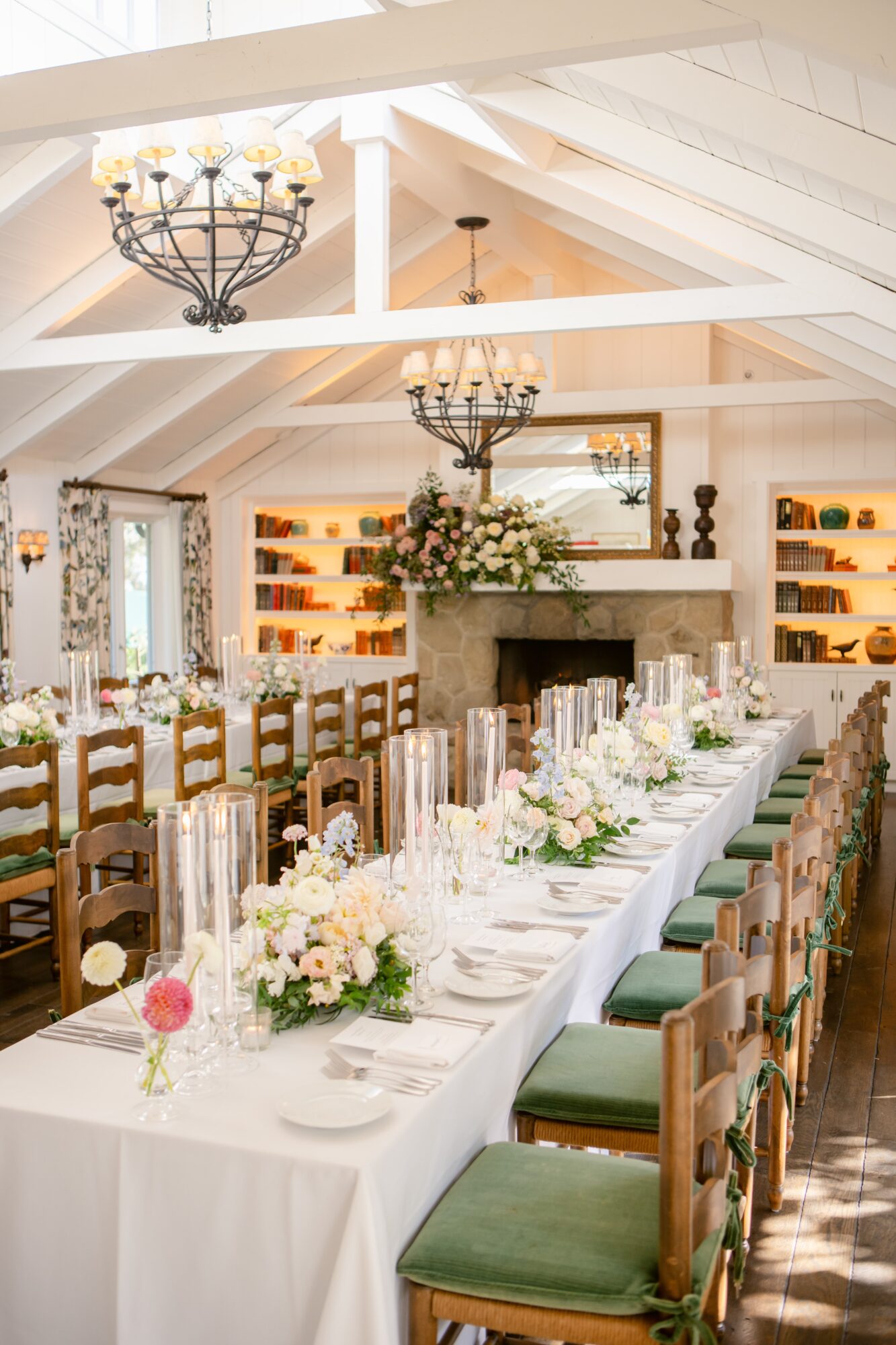 Long dining table with floral centerpieces and candles, set in a bright room with high ceilings and chandeliers.