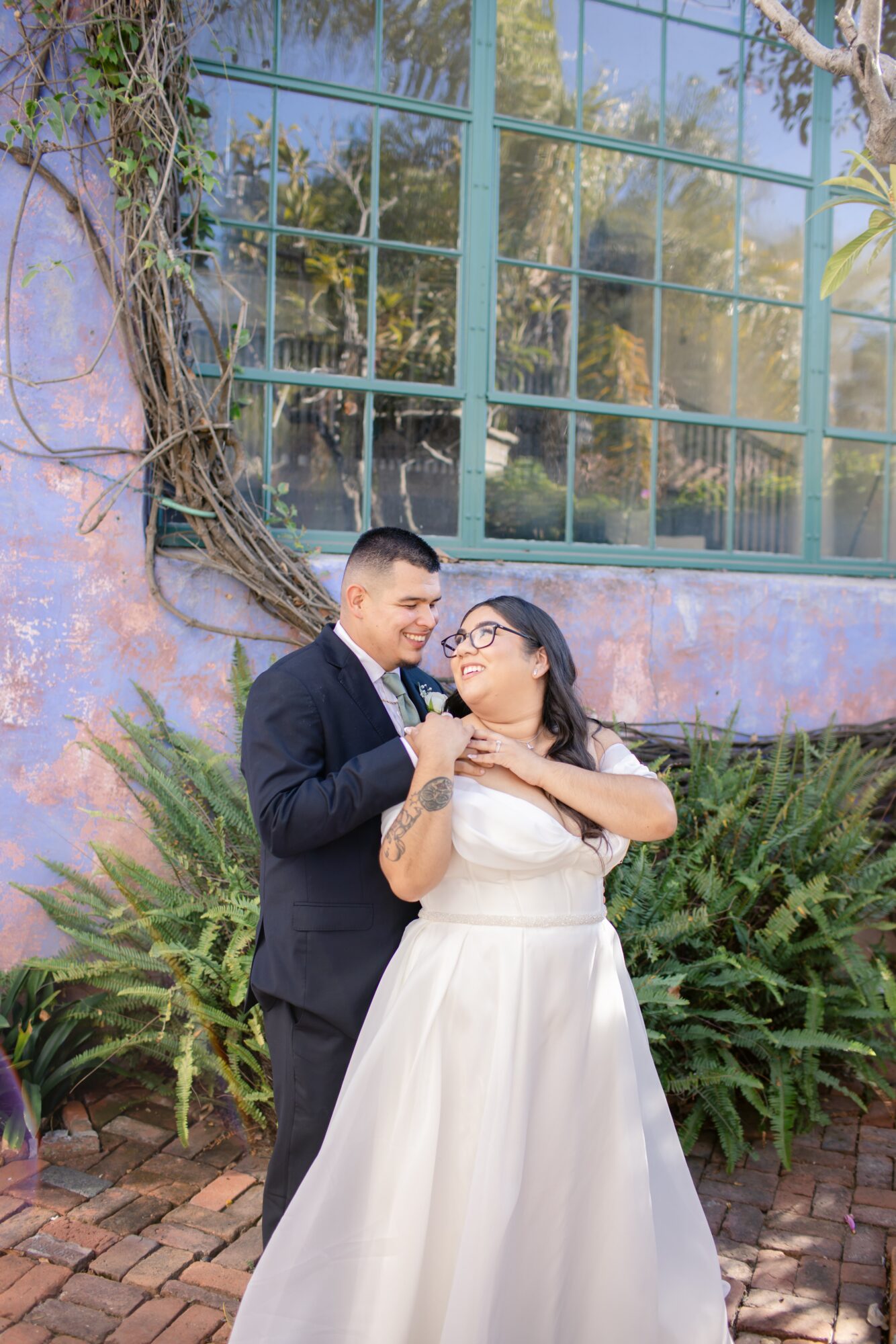 A couple dressed in formal attire standing outdoors, smiling and looking at each other, with greenery and a large window behind them.
