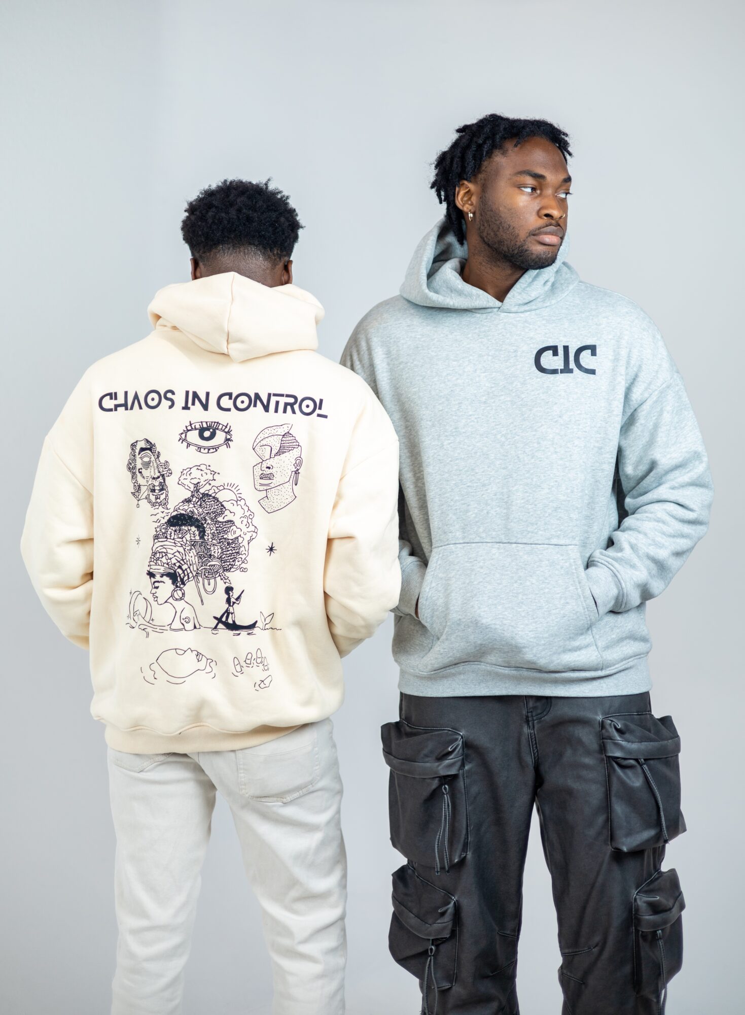 Two young men stand against a plain background, one facing away and the other looking to the side, wearing hoodies and cargo pants.