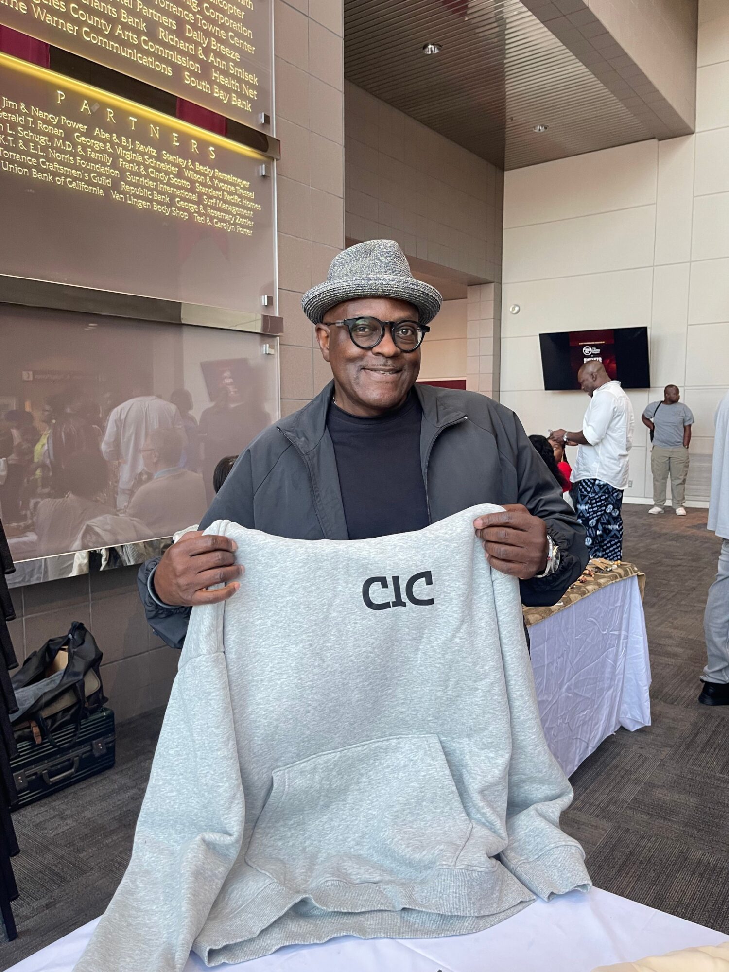 Person wearing glasses and a hat holding a large gray hoodie with 'CLC' text, standing indoors near a counter.