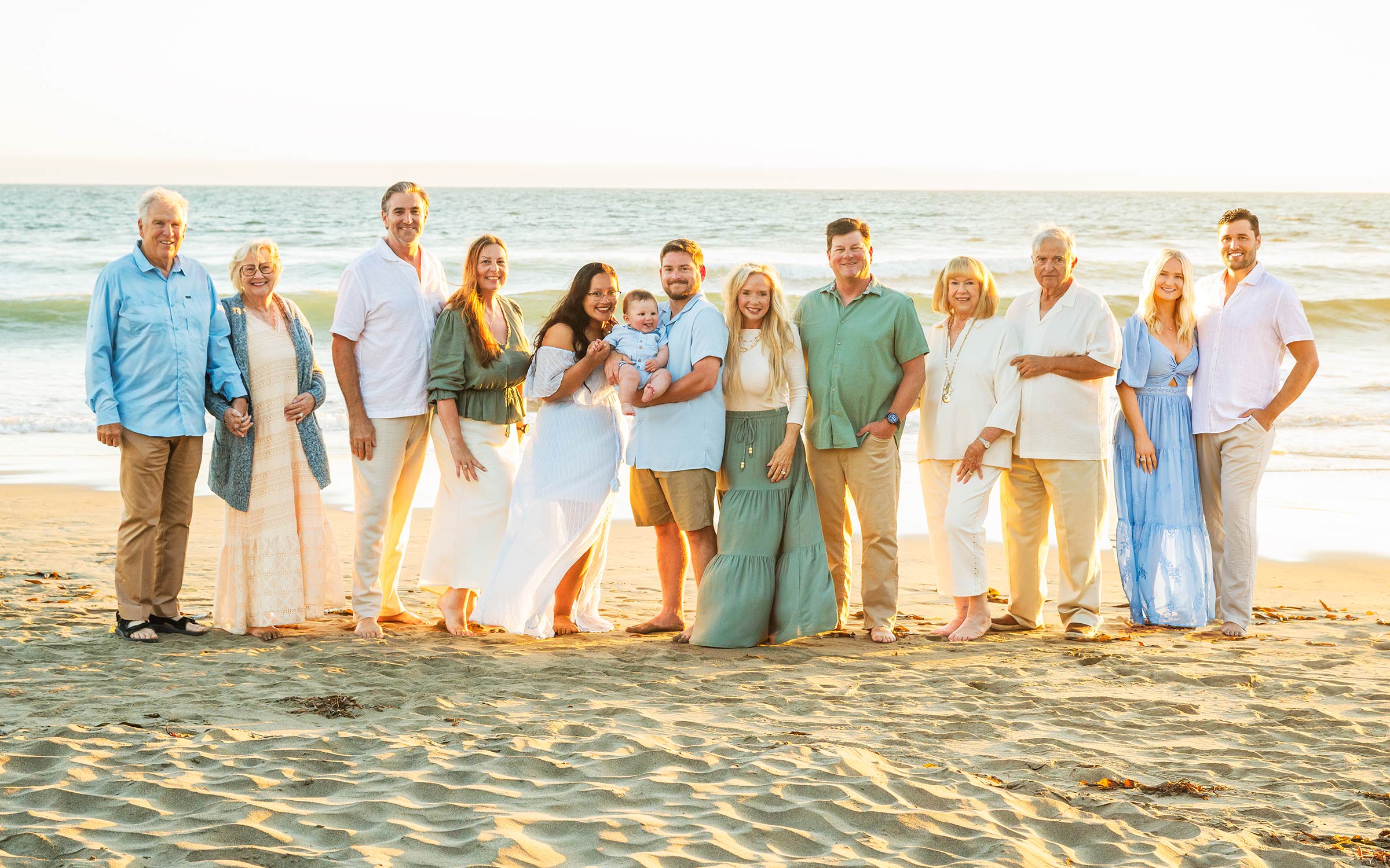 Group of people standing on a beach, smiling, with ocean and sky in background, some holding hands or with arms around each other.