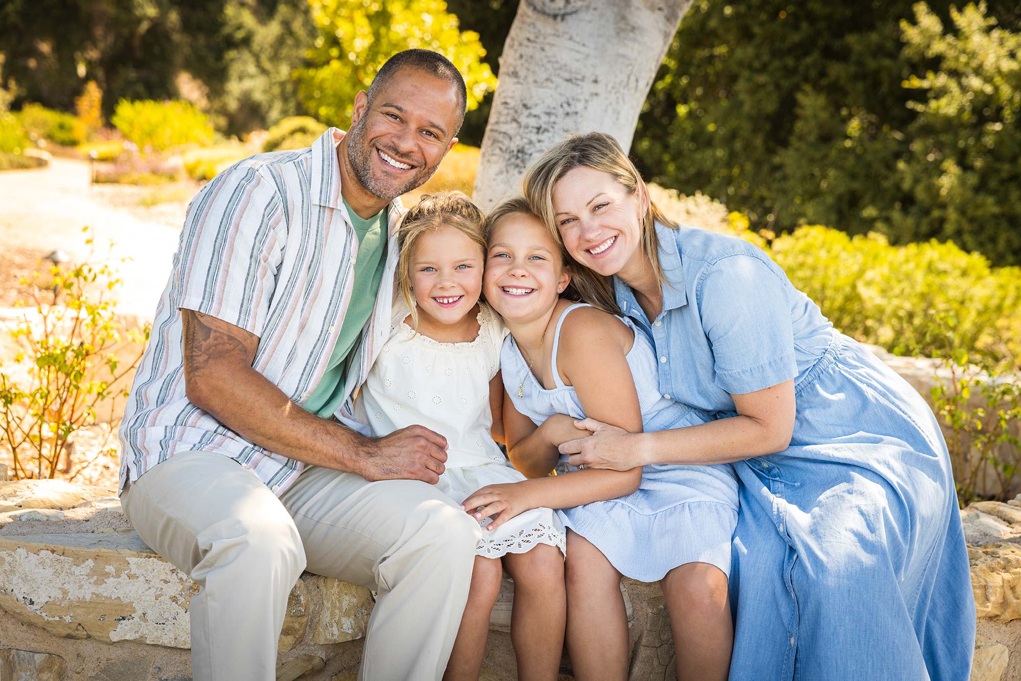 Family of four sitting outdoors on a stone bench, smiling, with trees and yellow flowers in background.