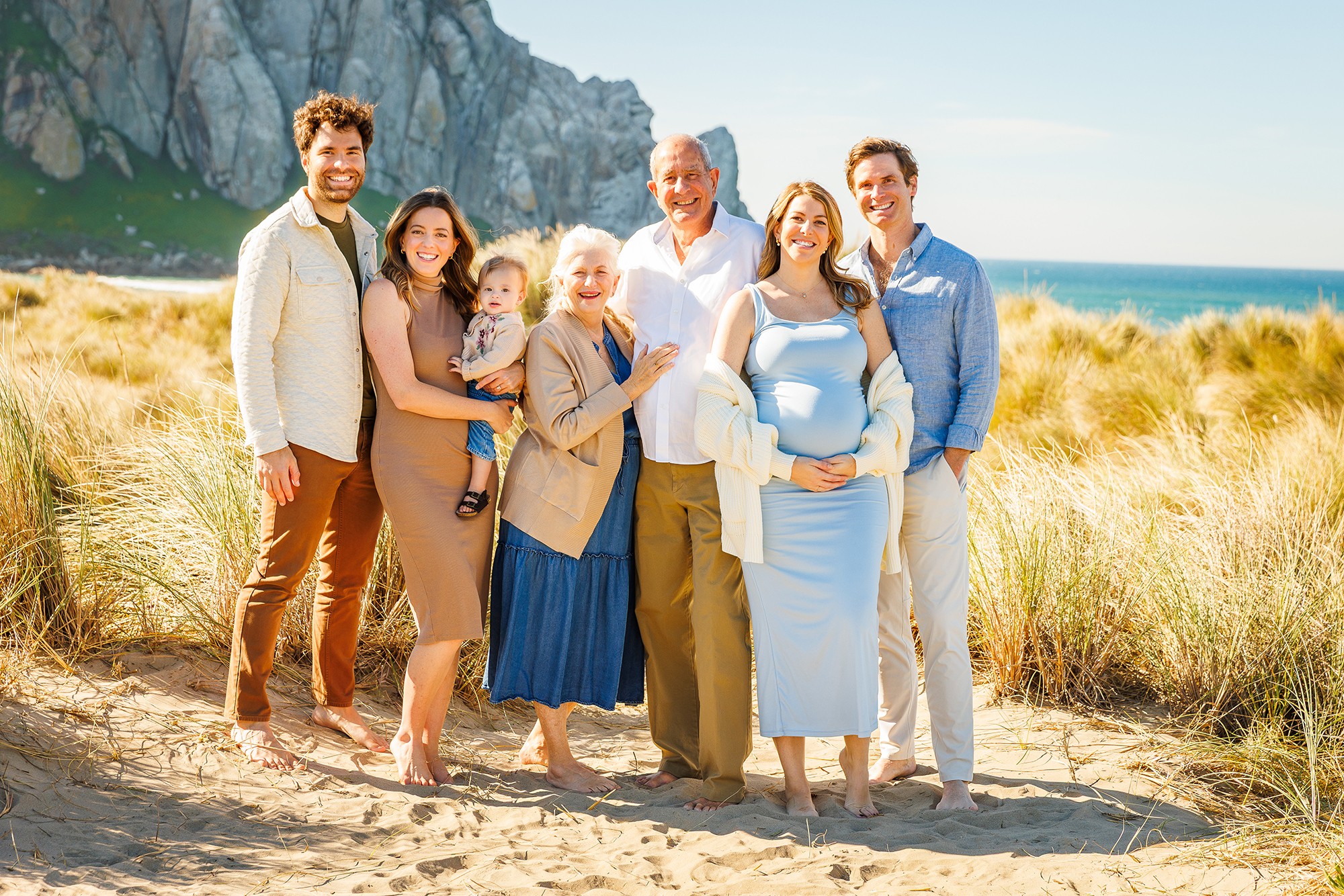 Group of seven people standing on a sandy beach with grassy dunes and cliffs in the background, smiling at the camera.