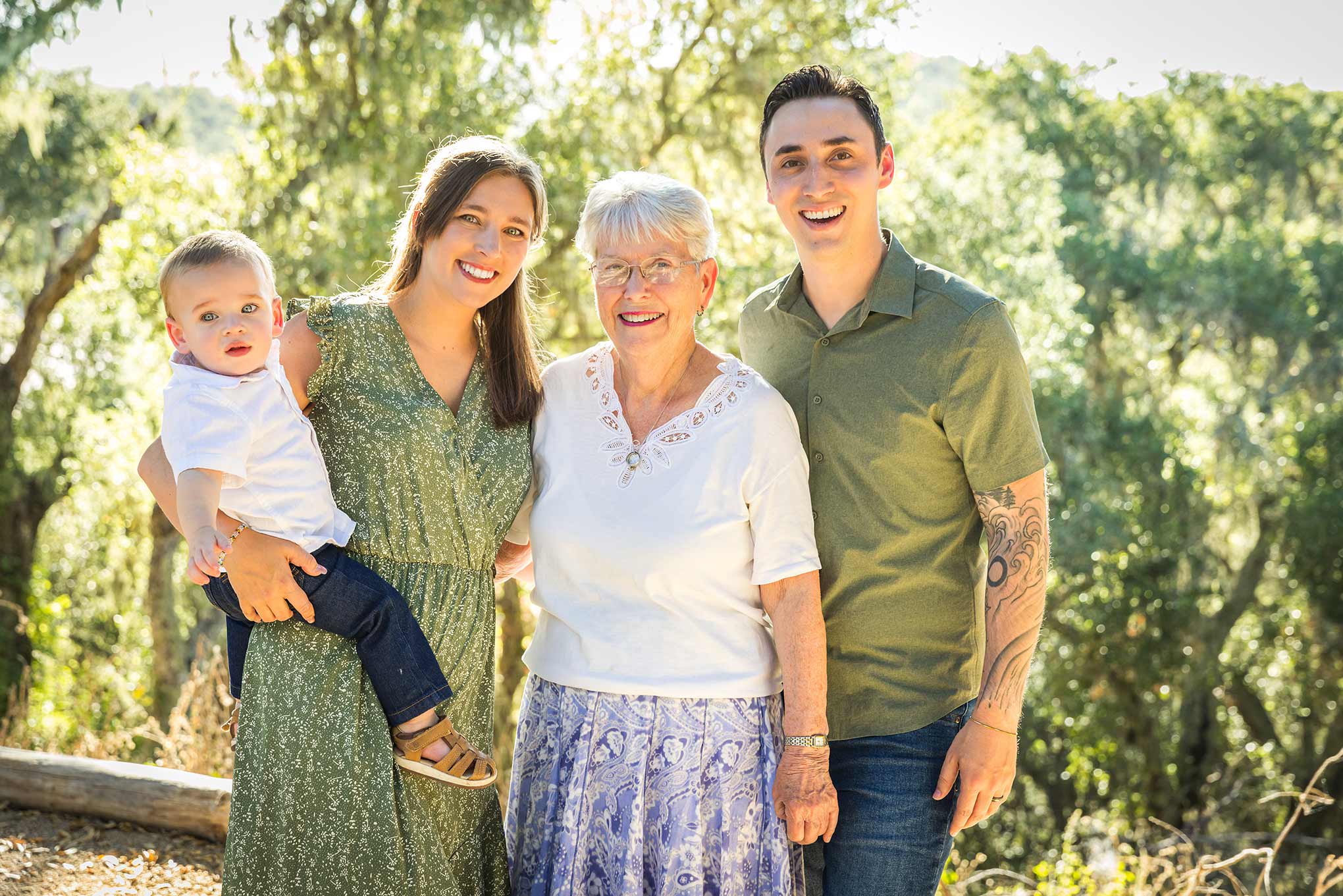 Four people, including a baby, standing outdoors in sunlight with trees in the background, smiling at the camera.