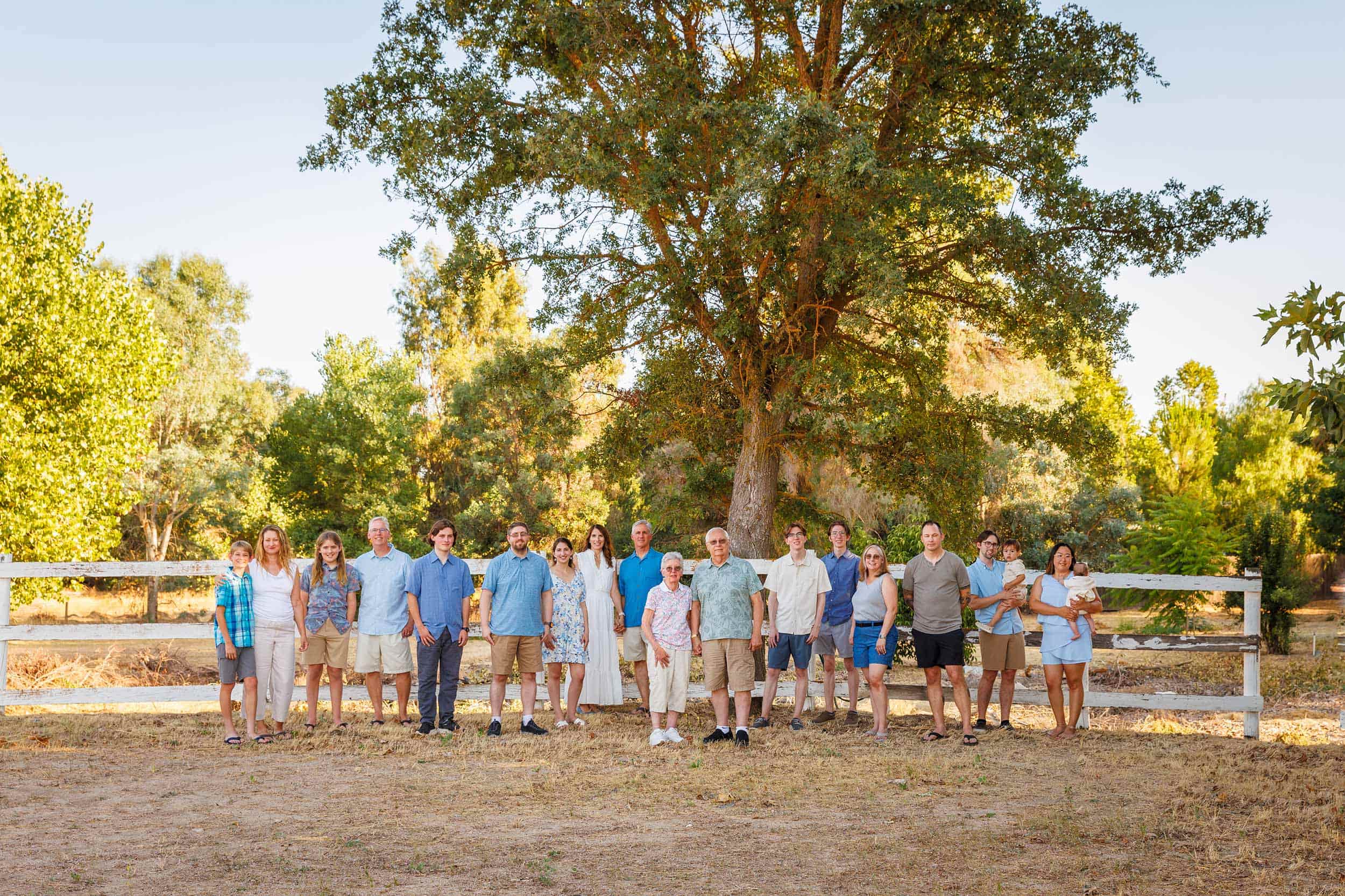 Group of people standing outdoors in front of a large tree and a wooden fence, with green trees and blue sky in background.