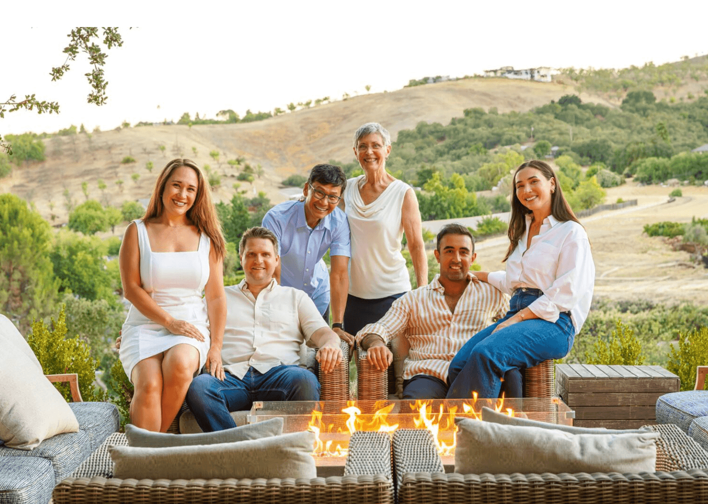 Group of six people outdoors near a fire pit, smiling, with hills and trees in the background.