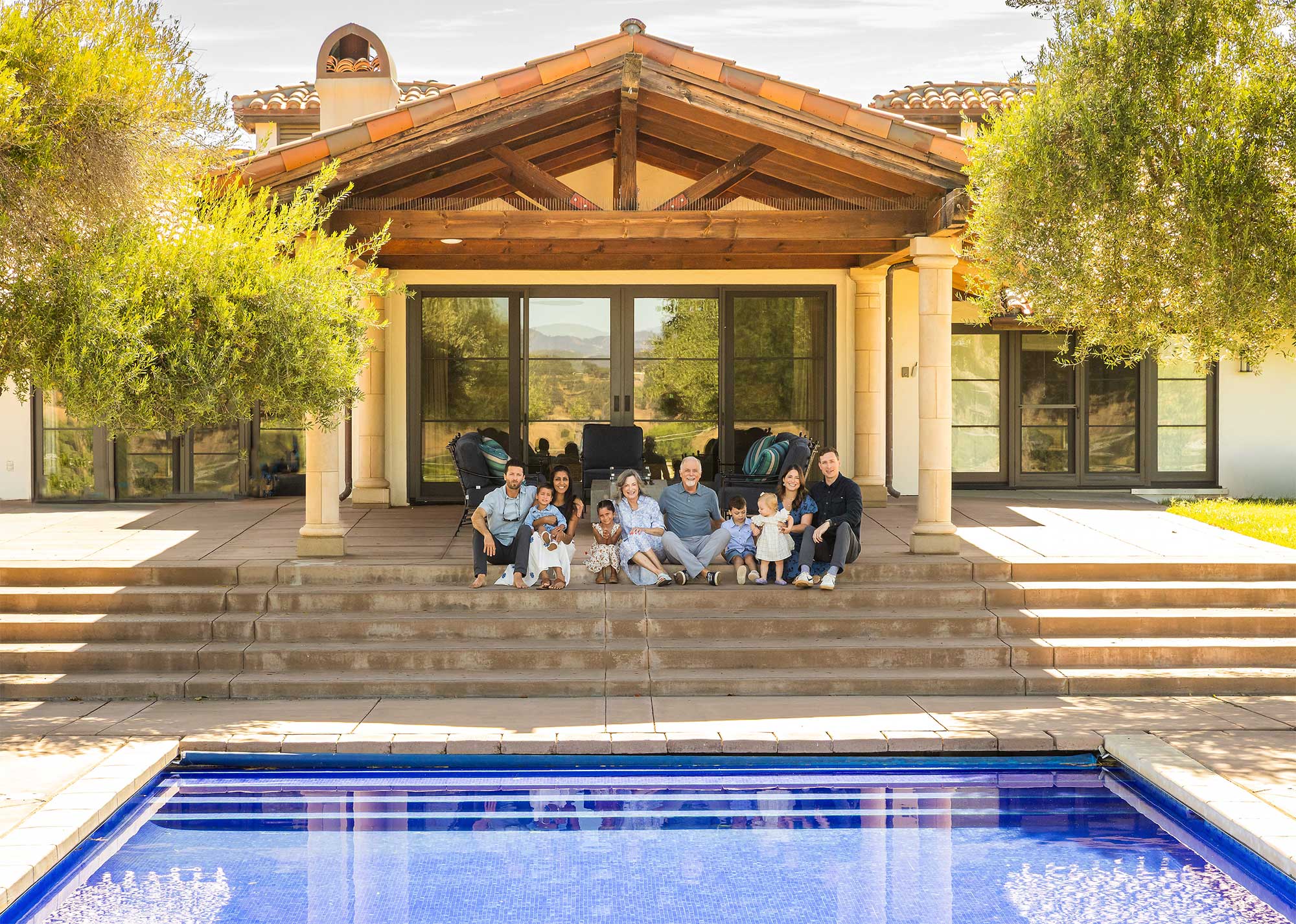 Group of people sitting on steps in front of a house with a pool in the foreground, trees on sides, and a wooden roof structure.
