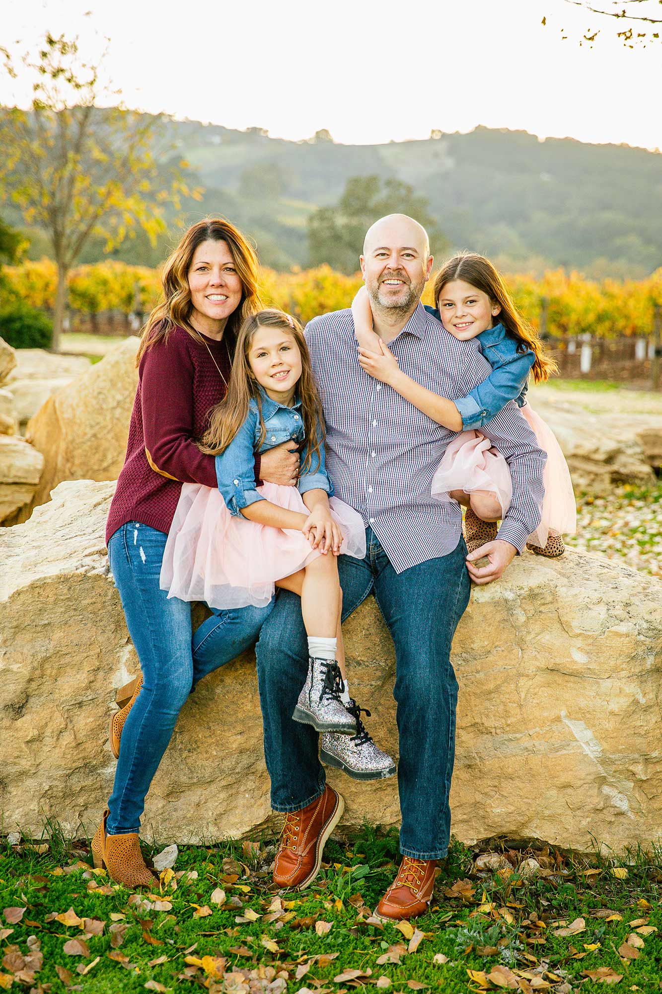 Family of four outdoors, smiling, with trees and hills in background, standing on a stone wall, autumn setting