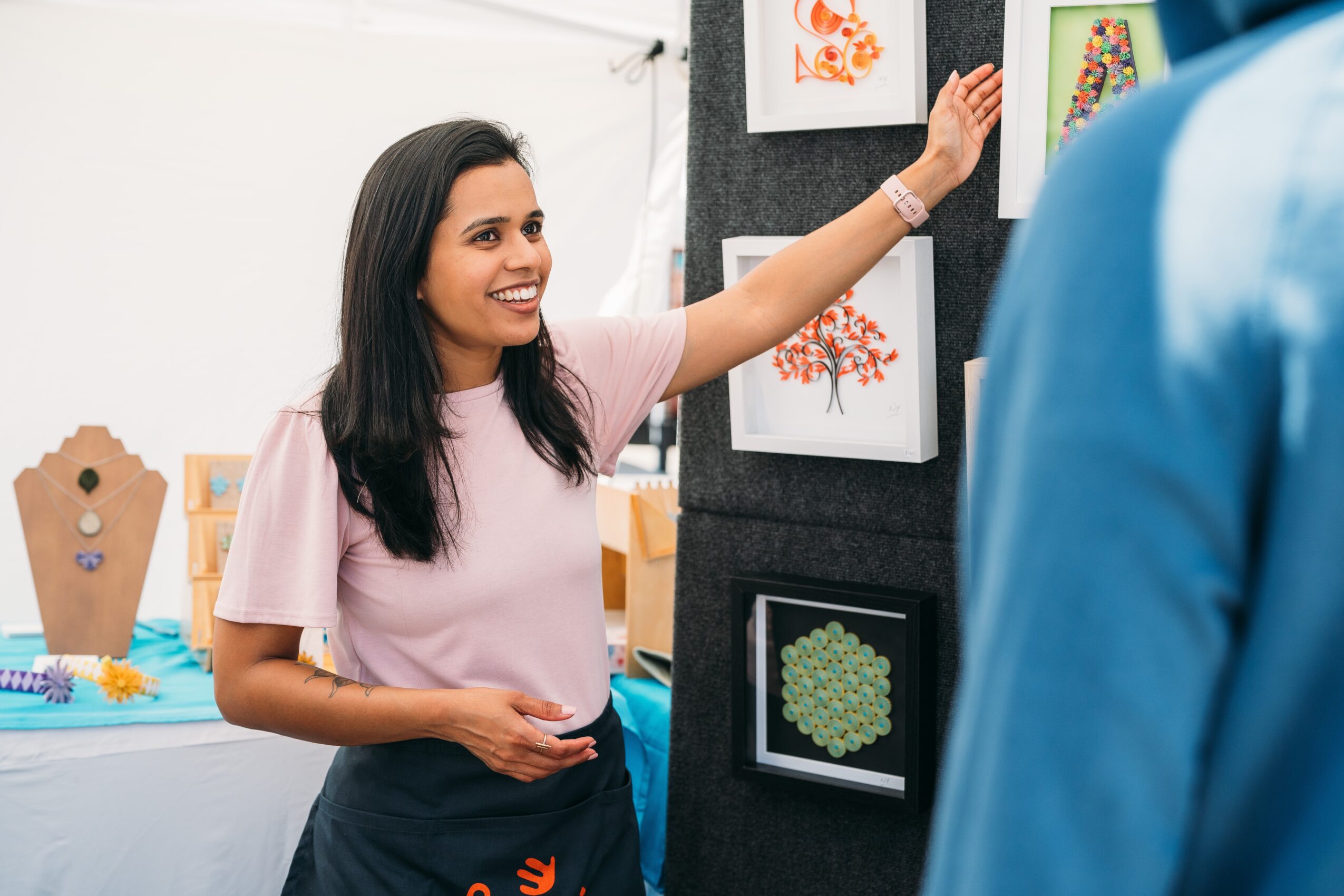 Smiling woman in pink shirt showing artwork to a person in blue, art display on wall behind her.