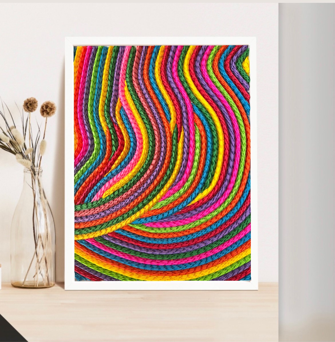 Colorful abstract yarn pattern with wavy lines and vibrant colors, framed and displayed on a wall, with a vase of dried flowers nearby.